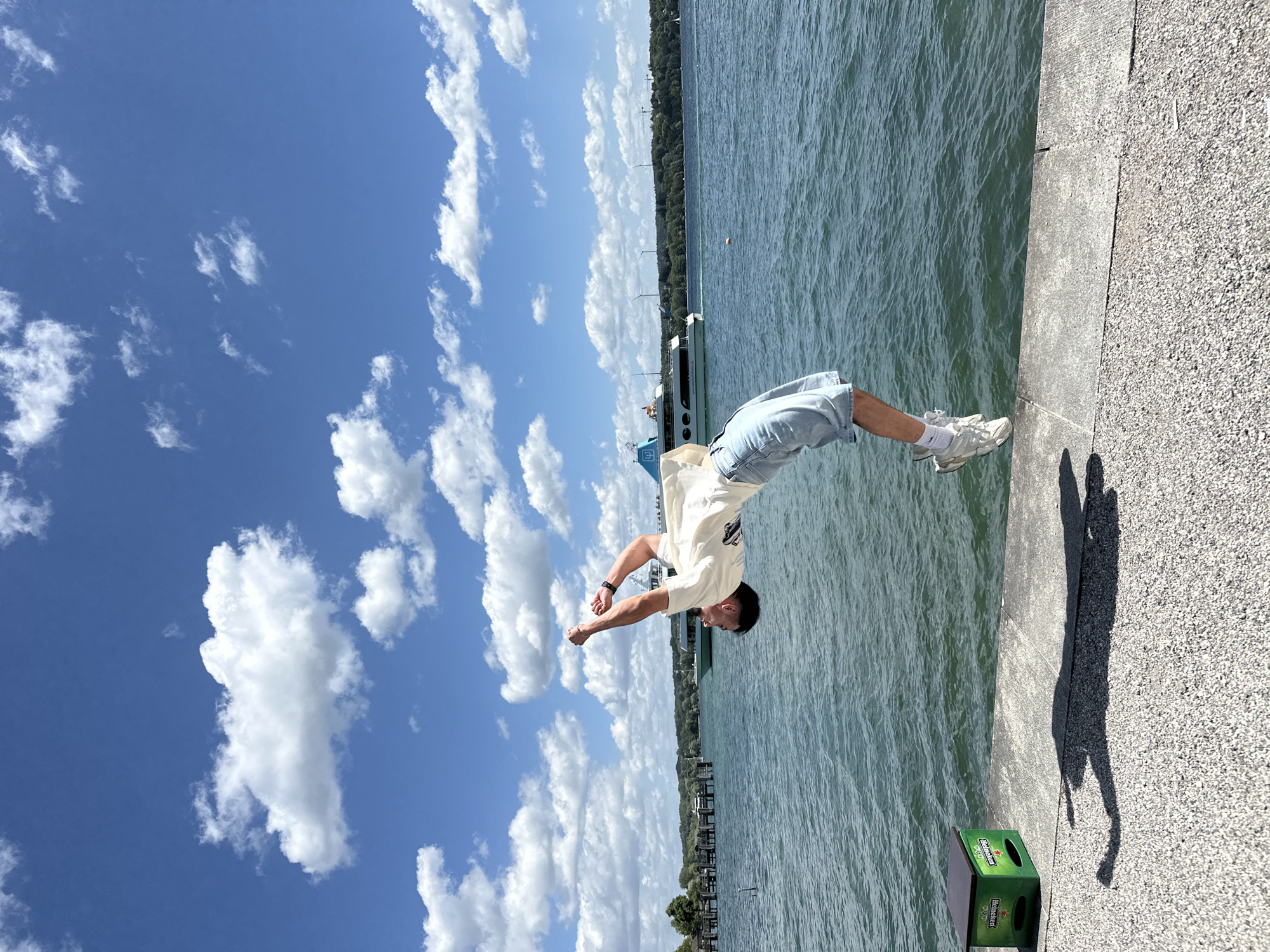 A young man is captured mid-air performing a backflip on a concrete embankment next to Lake Starnberg in Starnberg, Germany. He is wearing a light-colored t-shirt and denim shorts. The lake stretches out behind him, with a large white and blue boat visible in the distance, along with a tree-lined shore and several wind turbines. The sky above is a vibrant blue, dotted with fluffy white clouds. In the foreground, to the left, is a green Heineken beer crate. The man's shadow is cast on the ground, also appearing to be in motion from his flip.