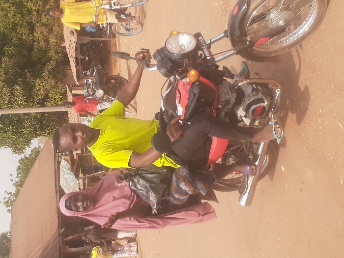 The scene depicts an unpaved street in Gwadanye, Nigeria. A man and a woman are positioned on a red and black motorcycle. The man, wearing a yellow t-shirt, dark trousers, and light blue flip-flops, sits in the driver's seat with his left hand on the handlebar, looking towards the viewer. The woman, a passenger, wears a pink hijab, a dark garment, and purple flip-flops. She holds a black plastic bag and gestures with her right hand, her mouth open, while also looking towards the viewer. The motorcycle features a chrome exhaust and the text "YOUNG MAN" visible on the front headlight assembly. The ground surface is reddish-brown dirt. In the background, multiple individuals are present, including a man in a red shirt walking to the right of center, and a person on a bicycle to the far right. Structures with corrugated metal roofs and green trees are visible, along with a utility pole in the mid-ground.