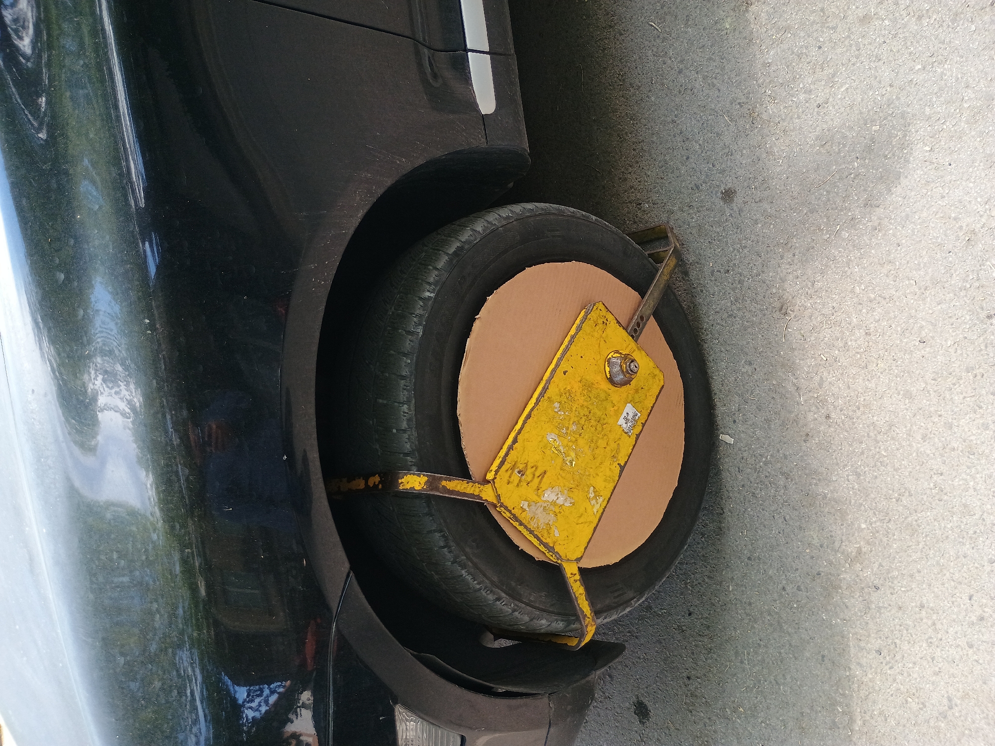 A black vehicle has a yellow wheel clamp attached to its front tire on a city street in Unknown, Unknown. A piece of cardboard is positioned inside the clamp, covering the tire. The street appears to be paved with concrete.