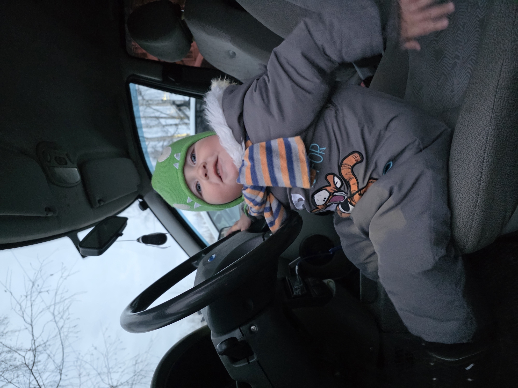 A young child, likely a toddler, sits in the driver's seat of a vehicle. The child wears a green hat with spikes, a grey puffy jacket with a cartoon tiger, and a striped scarf in shades of orange, blue, and grey. The child is holding the steering wheel. The vehicle's interior is grey. Outside, through the windshield, bare trees are visible against a grey sky. Based on the location details, this is likely a cold-weather environment in Chaikovsky, Russia. The photo appears to have been taken from an angle, possibly from the passenger seat or slightly above the driver's seat.
