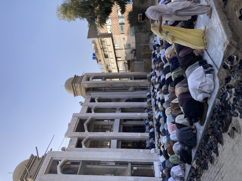 The scene depicts a large outdoor congregational prayer in Quetta City Tehsil, Pakistan. Approximately 100 to 150 men are observed prostrating (performing sajdah) on an elevated concrete platform. Their foreheads, noses, palms, knees, and toes are in contact with the ground, covered by individual prayer mats or small rugs. The individuals are dressed in various colored traditional attire, primarily shalwar kameez. In the immediate foreground, a substantial collection of shoes and sandals is neatly arranged along the perimeter of the prayer platform. To the far right, one man, clad in a light-colored shalwar kameez and a prayer cap, stands, holding and adjusting a large yellow fabric item, possibly a prayer rug.
The background features a multi-story mosque structure, seemingly under construction or recently completed, characterized by exposed concrete elements, arched window openings, and two domed minaret-like towers. Clothes are visible hanging on a balcony railing on the left side of the mosque. Satellite dishes are mounted on the upper sections of the building. Beyond the mosque and to the right, urban buildings are visible under a clear blue sky; one has multiple blue water tanks on its roof, and another features a patterned facade with a partially visible "WELCOME" sign. A green tree stands on the far right. The ground immediately beyond the prayer platform appears to be unpaved or dirt.