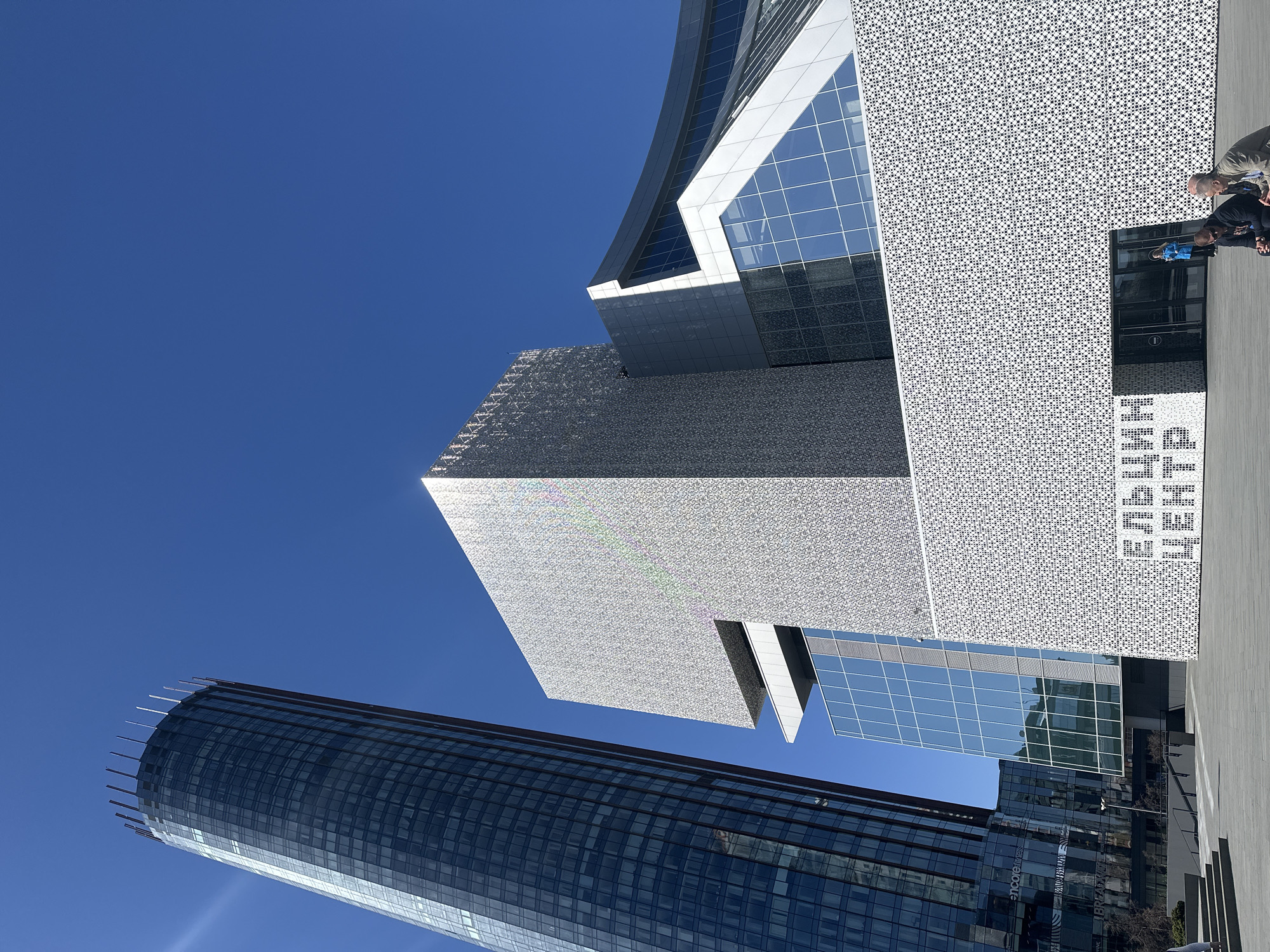The image shows a vertical shot of three buildings in Yekaterinburg, Russia, against a clear blue sky. In the foreground is the Iset Tower, a cylindrical skyscraper with a glass facade. Above and behind the tower is the Yeltsin Center, a modern building with a mosaic-patterned facade and glass sections. Two people are visible in front of the Yeltsin Center entrance. The ground appears to be paved with light-colored stones. The overall scene conveys a sense of modern urban architecture.