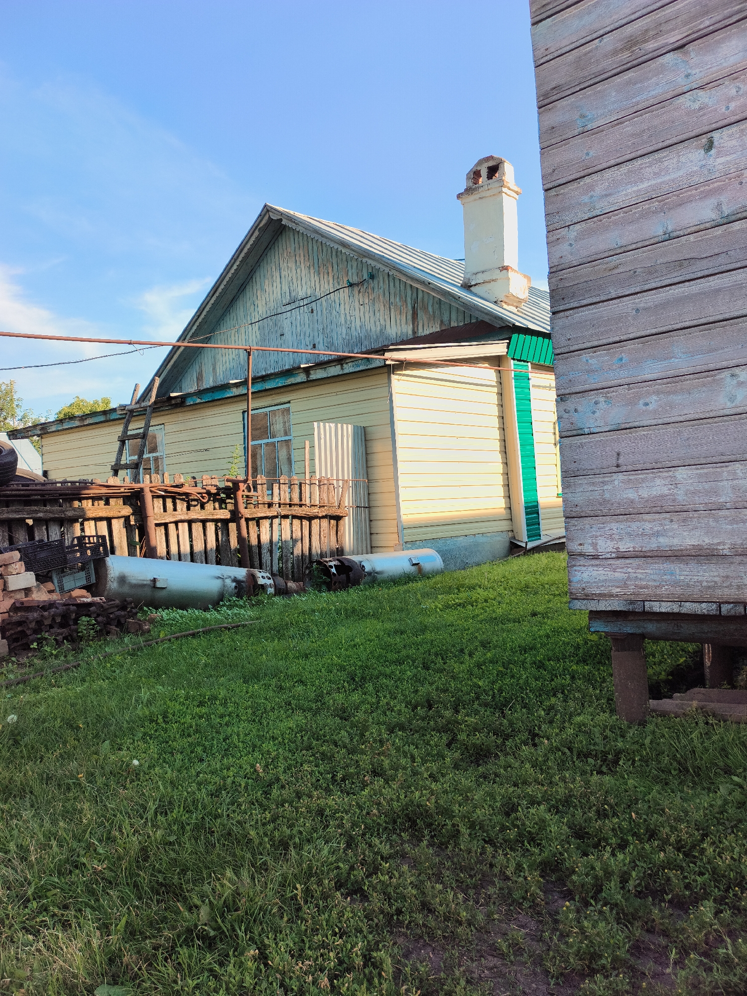 This is a medium-shot, eye-level, outdoor, full-color photograph of a rural dwelling in the village of Al'kino, Russia, on a clear day with blue sky and scattered clouds. The scene captures a section of a light yellow wooden house with a light blue weathered gable end, a silver metal roof, and a white brick chimney with visible openings at the top. A wooden ladder leans against the house. In front of the house is a dilapidated wooden fence with various metal objects, including what appear to be large cylindrical tanks, and piles of bricks and tires. The foreground is covered in lush green grass. To the right of the frame, a portion of a weathered wooden structure, possibly an outbuilding, stands elevated on concrete blocks.