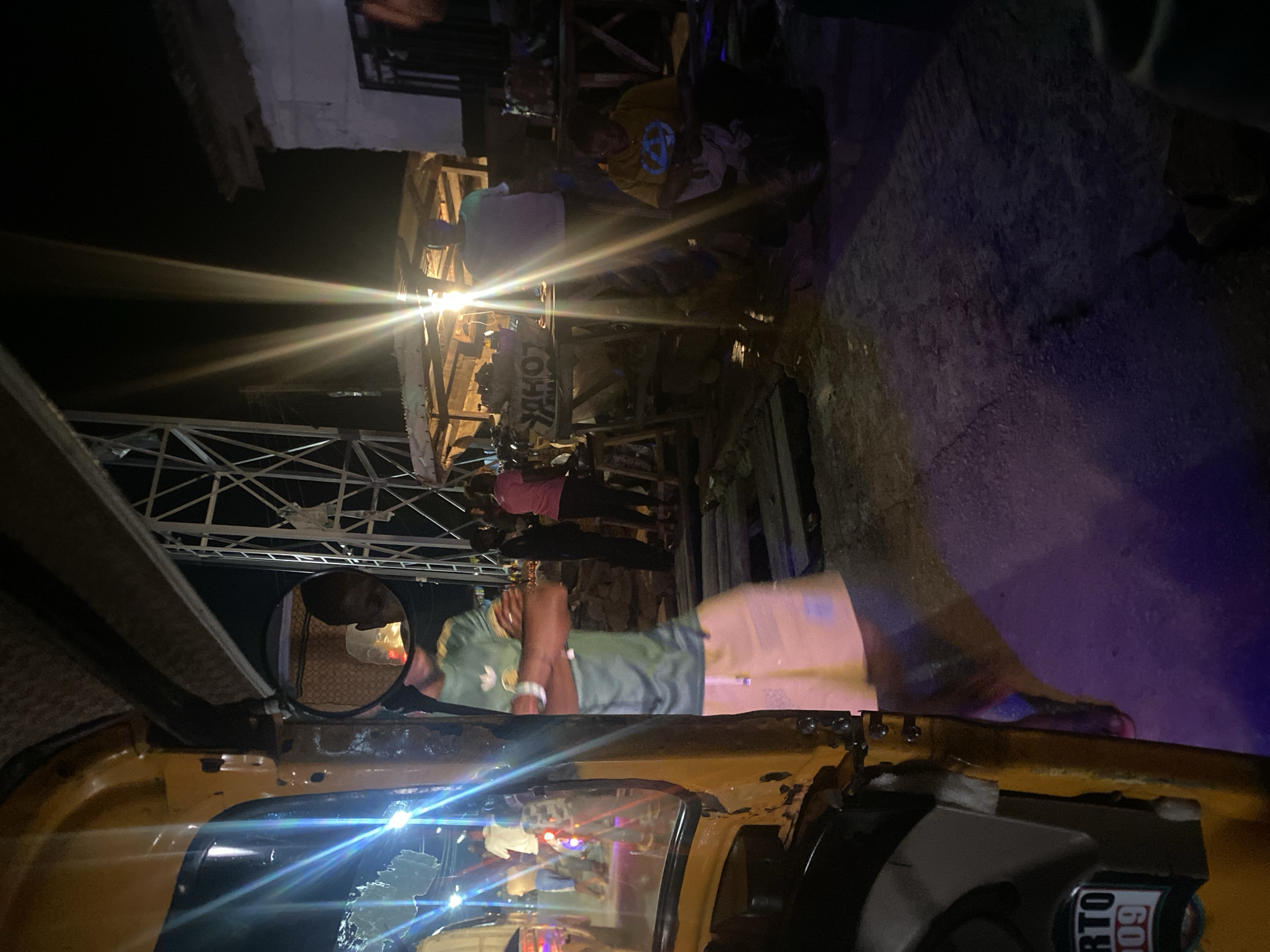 The image captures a nighttime scene in Jos, Nigeria, with artificial lights creating strong glare and dramatic shadows. In the foreground, the left side of a yellow auto-rickshaw is visible, its side panel showing a sticker with text "RTO 1009". A side-view mirror reflects a person's head and shoulders.
The background depicts a bustling, somewhat chaotic environment, possibly a street market or gathering. Several individuals are present. A person in a green t-shirt and light-colored shorts stands with arms crossed, facing away from the camera. Behind them, a person in a pink shirt and another in a dark shirt and pants are also visible. Further back, two figures stand near a wooden structure that appears to be a stall or makeshift shelter, illuminated by bright lights. One person in a white t-shirt stands, while another in a yellow t-shirt is seated on the ground, looking at something in their hands. A large metal structure, possibly part of a construction site or temporary event setup, stands prominently in the middle ground, its framework illuminated by lights. The ground is a mix of dark, textured pavement and areas illuminated by colored lights, particularly a strong purple hue on the right side. The overall impression is of an active outdoor space at night.