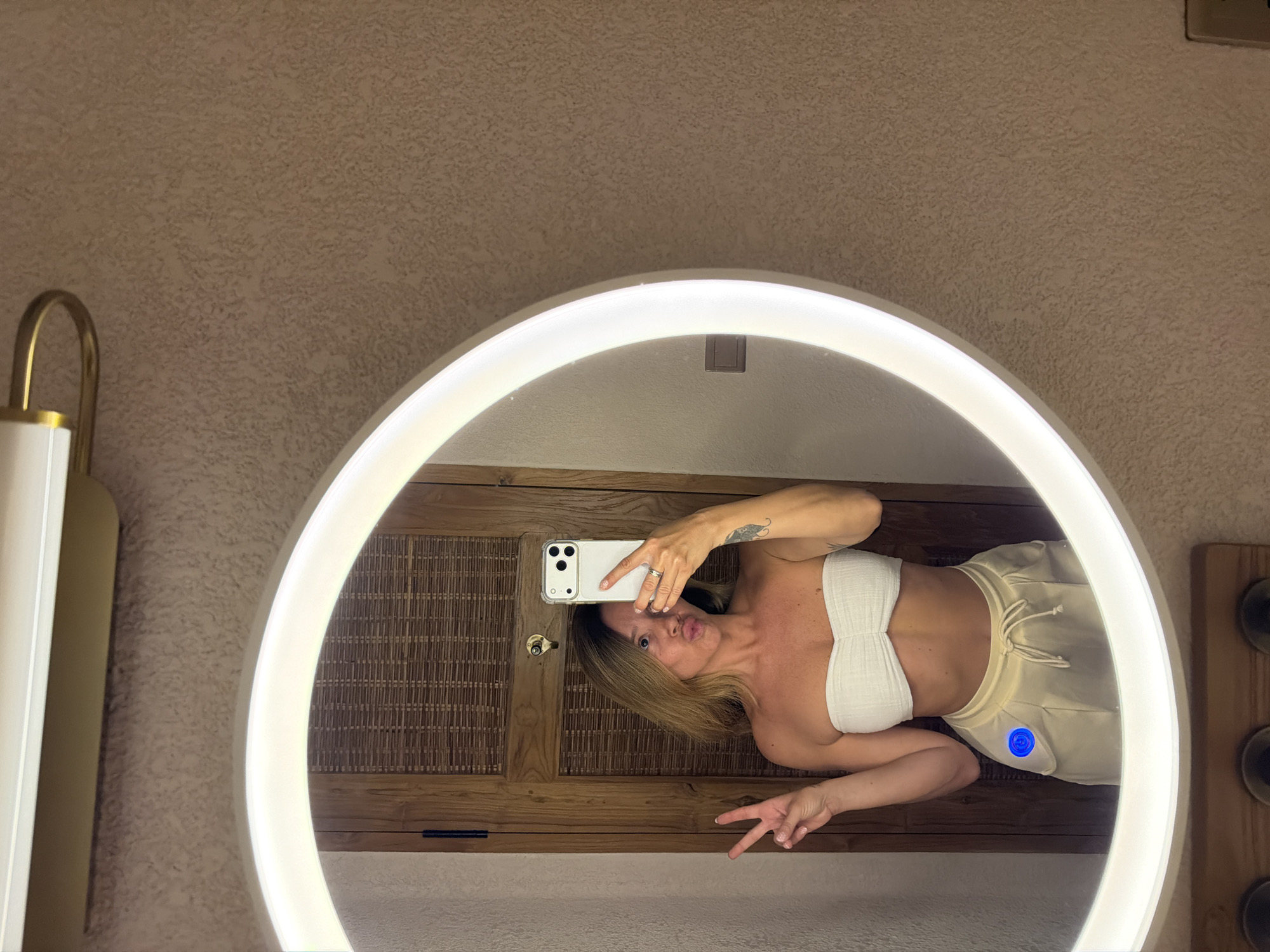 This image captures a mirror selfie taken by a woman in an indoor setting, likely a bathroom or dressing area in Mengwi, Indonesia. The original image is rotated 90 degrees counter-clockwise.
The central subject is the reflection of a woman in a large, circular mirror that is brightly illuminated by an integrated LED light strip around its circumference. The woman has blonde or light brown hair and is wearing a white, textured bandeau top and light-colored (beige or cream) drawstring shorts. A tattoo is visible on her right upper arm. She is holding a white smartphone, likely an iPhone, with her left hand, and appears to be taking the selfie. Her expression is playful, with puckered lips (a "duck face"), and her right hand is making a peace sign. A ring is visible on her left hand, and her fingernails have a glittery polish.
Behind the woman's reflection, a wooden surface with a distinct slatted or woven pattern is visible, suggesting a cabinet or wall panel. A brass keyhole is present on this wooden surface near where she holds her phone. A unique detail is a circular blue light or button located on the outer right thigh of her shorts.
The wall surrounding the mirror has a textured, light brown finish. To the left of the mirror, a modern, gold-toned curved fixture, possibly part of a light or faucet, is partially visible. The overall lighting is bright, primarily from the mirror's LEDs. The scene conveys a casual and lighthearted mood.