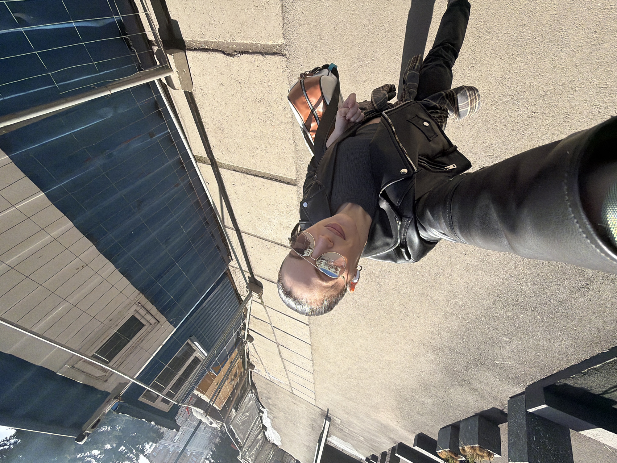 A person takes a selfie outdoors on a sunny day. The person is wearing a black leather jacket, black shirt, dark pants, and round sunglasses, and carrying a bag. They are standing on a gray paved surface. In the background is a building with blue and white paneling, partially obscured by a metal fence. In the bottom left corner, there is greenery, possibly trees. To the bottom right, there are dark grey rectangular posts. The location is Perm, Russia.
