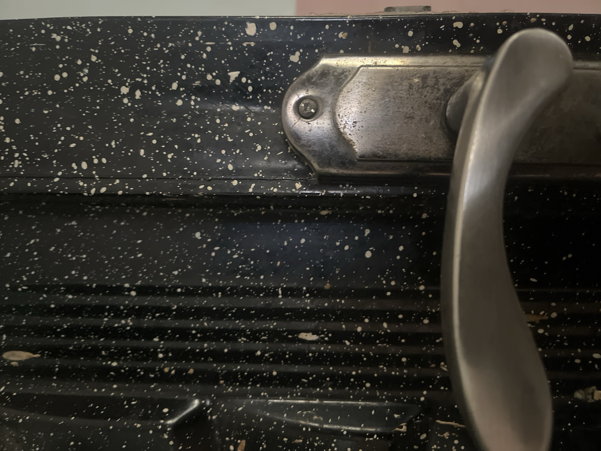 The image is a close-up, horizontal shot of a black, speckled door handle and part of the door it is attached to. The door has a textured, ribbed surface with many small white speckles scattered across it, as well as a few larger, irregular brown marks. A silver-colored door handle is prominently featured in the foreground, extending from the right side of the frame. Above the handle, a metal escutcheon plate is visible, secured by a screw. The scene appears to be indoors, given the lighting and the typical appearance of a door. There are no people or animals present in the image. The background is out of focus and shows a light-colored surface. There is no information or visual context within the image to suggest its location in Jalingo, Nigeria.