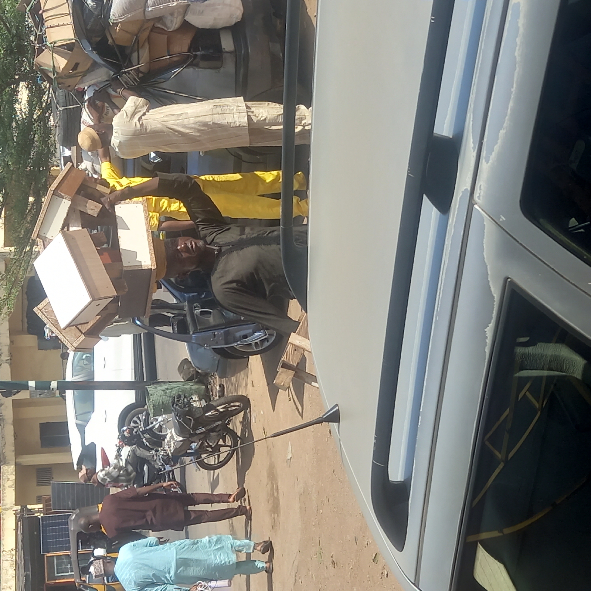 This photograph captures a bustling street scene in Dadi Riba, Nigeria, likely during the daytime under clear weather conditions. The primary focus is on a dark-colored vehicle, possibly a car or small truck, heavily overloaded with various goods secured to its roof and rear. The cargo includes numerous cardboard boxes, large sacks, and what appears to be wooden furniture or frames, stacked high above the vehicle's roofline.
Several individuals are engaged around this loaded vehicle. One man, dressed in a yellow long-sleeved shirt and dark trousers, is prominently featured, holding or carrying a large wooden structure with white panels, indicating he is actively involved in the loading or unloading process. Another man in light-colored, striped traditional attire and a matching cap is also near the overloaded vehicle, observing or assisting. A third man in a dark grey or black outfit is partially visible, contributing to the activity.
In the midground and background, further street activity is visible. A light-colored car, possibly an SUV or van, occupies the right foreground, exhibiting some wear with peeling paint on its roof and side. Behind the central activity, a white car and two motorcycles are parked. Several other men are seen standing or walking; one is dressed in a brown traditional outfit, another in a light blue attire, and one is seated on a motorcycle. To the far left in the background, a small stand with several solar panels is visible, suggesting a local energy solution. The ground is a mix of dirt and sand, and simple building facades line the street. No discernible text is visible in the image.