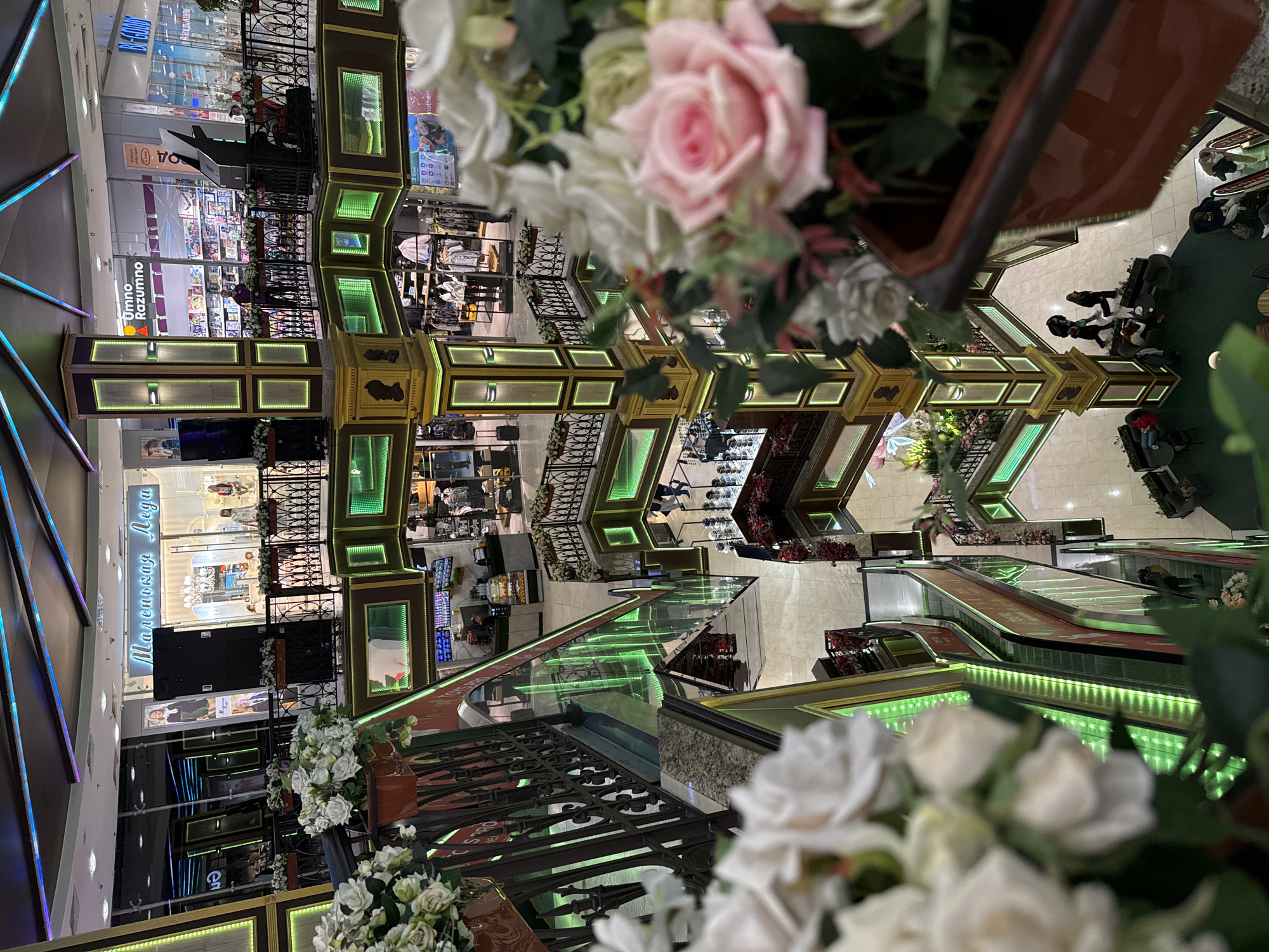 The image shows an interior view of a shopping mall in Moscow, Russia. The mall features multi-level floors with decorative barriers accented with faux flowers and green lighting. Several shops are visible, including one named "Umno Razumno" and another "Malenkaya Lyega". People can be seen walking around, and some are sitting at tables. Escalators are also visible. The view is partially obscured by a foreground arrangement of white and pink roses.