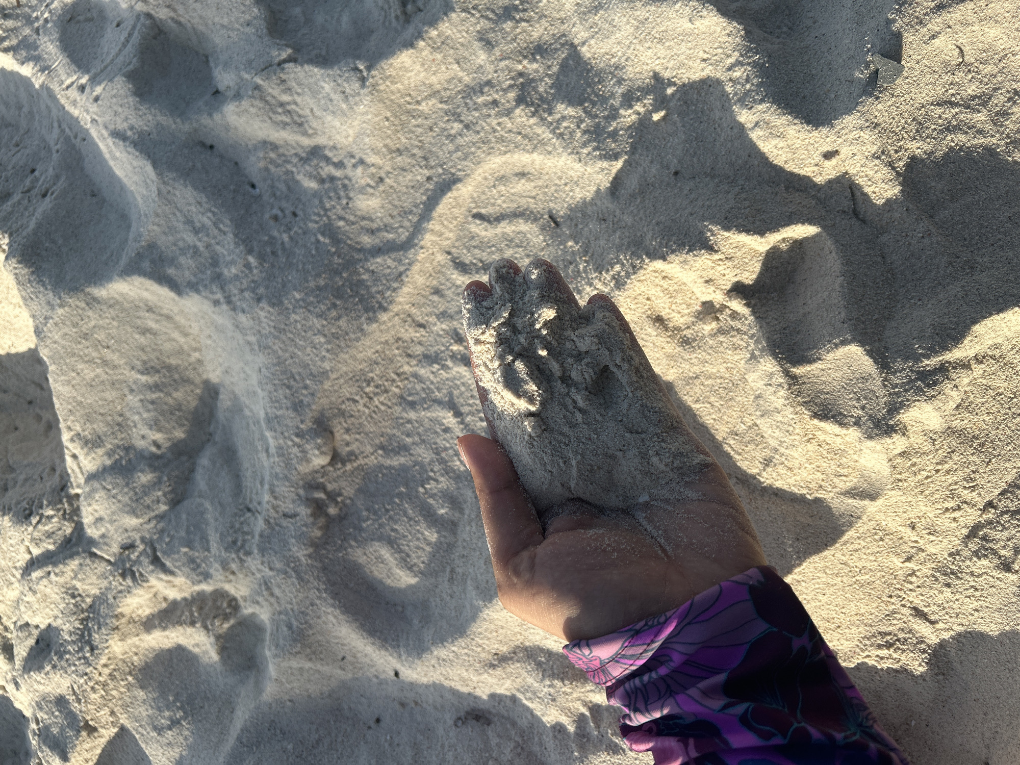 A close-up, overhead shot shows a person's hand holding a handful of light-colored sand. The hand is cupped, and the sand is slightly mounded within it. The person is wearing a purple rash guard with a floral pattern on the left arm, which is visible in the lower left quadrant of the image. The background consists of a vast expanse of rippled sand, likely a beach, with shadows cast by the uneven surface, indicating the presence of sunlight. The texture of the sand appears fine and soft. The overall lighting suggests it is either early morning or late afternoon. The scene is located on a sandy beach in A R A, Indonesia.