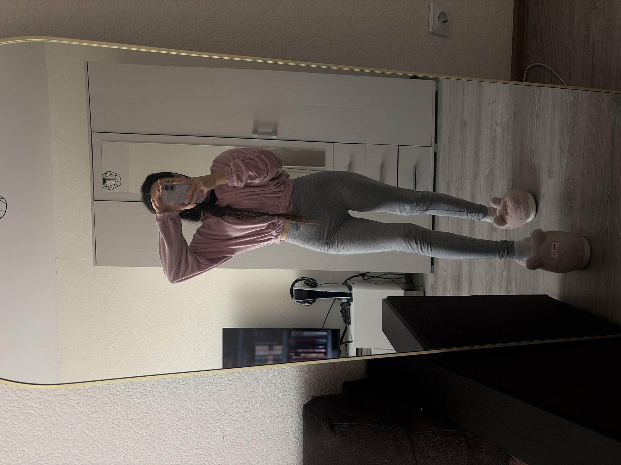 A person is reflected in a large mirror. The person is wearing a pink long-sleeve top and gray leggings, with plush bear-shaped slippers on their feet. They are holding a smartphone, presumably taking the photo of themselves in the mirror.
In the background, also visible in the mirror's reflection, is a white cabinet with silver drawer pulls, a TV stand with a TV, a PlayStation 5 with headphones, and dark wood furniture that blends into a dark sofa. The room is dimly lit.
There is no evidence of the location being in Saint Petersburg, Russia. There is no information to suggest that the location is in the city of Saint Petersburg, Russia. The surroundings do not reflect the architecture or cultural elements commonly associated with Saint Petersburg.
