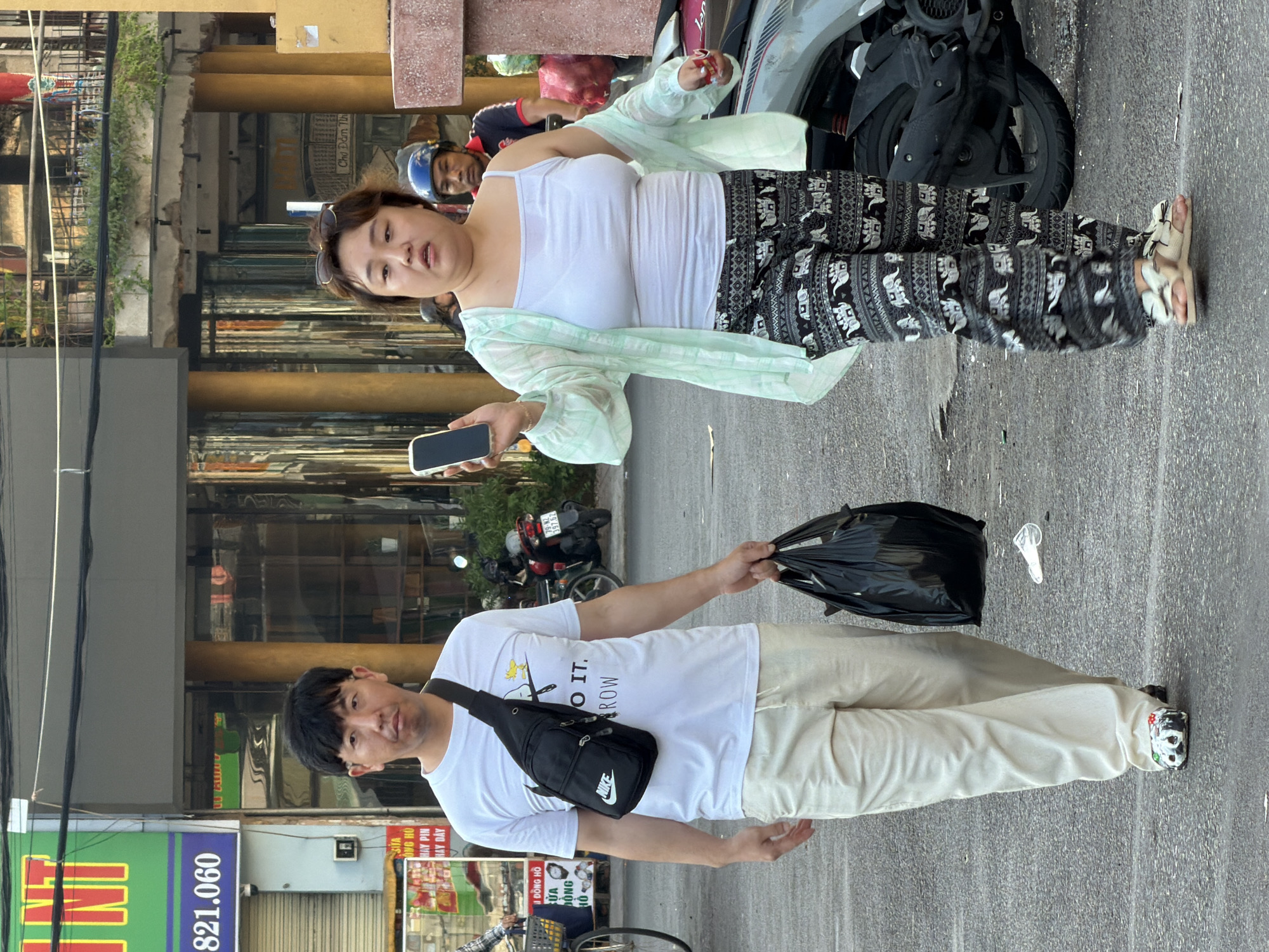 A man and a woman walk down a street in Nha Trang, Vietnam. The man, wearing a white t-shirt and light-colored pants, carries a black plastic bag in his right hand and a black crossbody bag over his left shoulder. The woman, in a white tank top and patterned pants, holds a smartphone in her right hand and has a light green shawl draped over her shoulders. They are walking on an asphalt road in front of buildings with glass storefronts and yellow pillars. A parked motorcycle is visible on the right side of the image.