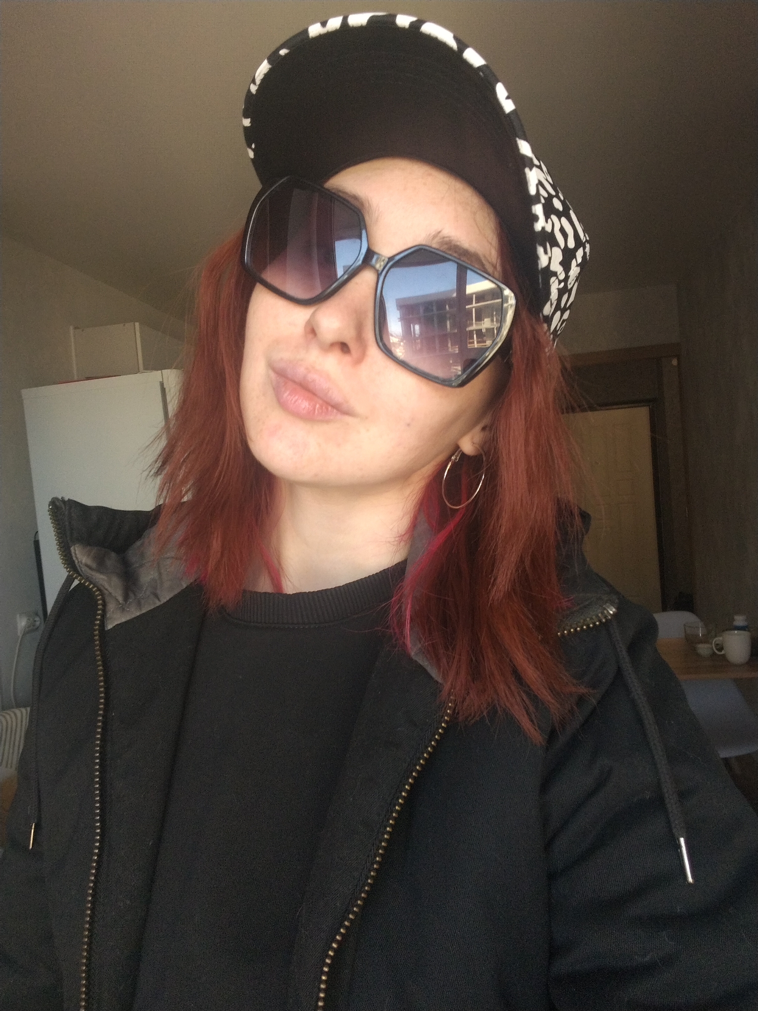 A female subject with red hair wearing a black and white patterned baseball cap and oversized black sunglasses is indoors. She is also wearing a black sweatshirt and a black jacket with the zipper partially zipped. A gold hoop earring is visible in her left ear. The background suggests an interior space, possibly an apartment or home, with a white refrigerator visible in the background left. A closed, medium-brown door with a beige wall and a glimpse of furniture, including a table with a white mug, suggests a dining or living area. Based on the available details, no explicit evidence confirms the location as Izhevsk, Russia.