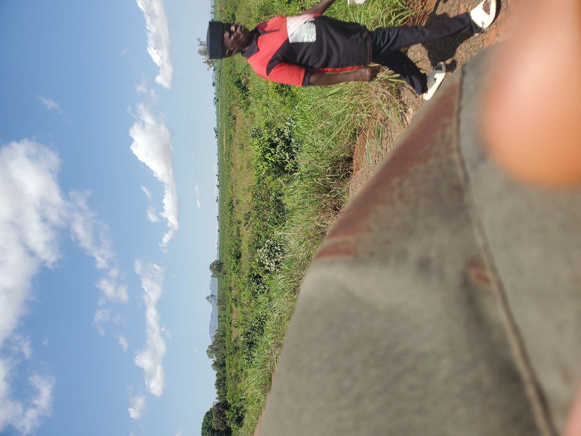 An elderly Black man wearing a black cap, a red and black polo shirt with a white emblem, black pants, and white sandals walks on a dirt path next to a field of green vegetation under a cloudy blue sky. In the foreground, a blurry, tan, curved object occupies the left side of the frame. The man is facing right, with his body angled slightly towards the viewer. The vegetation appears to be crops or wild growth, with some taller grasses and bushes interspersed. In the background, distant hills or mountains are visible on the horizon. The lighting suggests it is daytime, likely late morning or early afternoon.