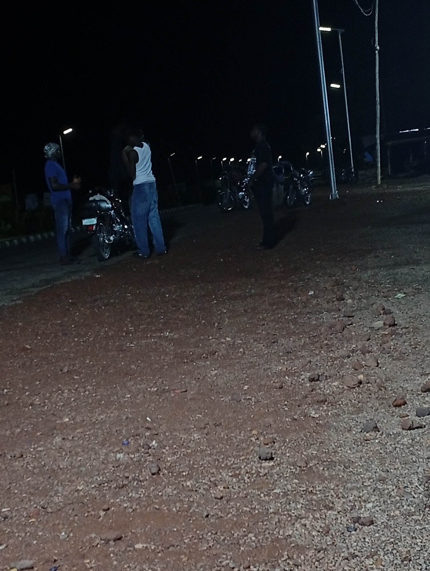 The image depicts a night scene in Adankolo, Nigeria. Three individuals are present near several motorcycles. One person wears a blue shirt and head covering, standing near a motorcycle, and another wears a white tank top and jeans. The third person is dressed in dark clothing. The ground is dirt and gravel. Streetlights illuminate the area.