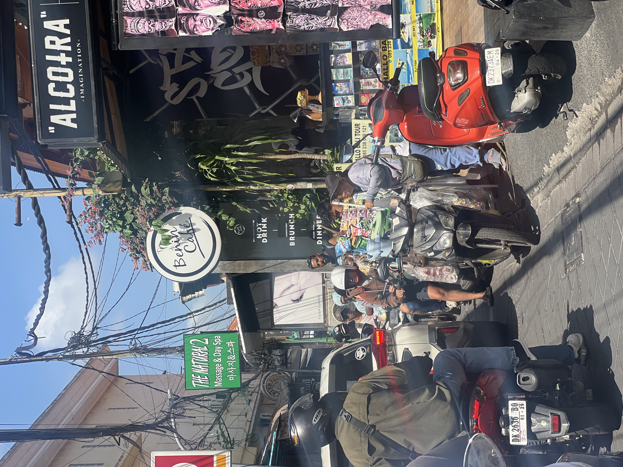 The image depicts a street scene in Kuta, Indonesia, characterized by heavy traffic, primarily composed of scooters and motorcycles. The foreground features a rider on a black scooter, his back to the camera, wearing a green jacket and blue jeans. To the right, a red Vespa scooter is parked, with its rear license plate visible, reading "DK 2373 AEM 04.29". Further back, a line of motorcycles and scooters stretches down the street, with several individuals wearing helmets.
On the right side of the street, a shop displays various items, including what appear to be posters or artwork of skulls and faces. Above the shop, a black sign with white lettering reads "\"ALCO+RA\" -IMAGINATION-". A circular sign with "Benin Cafe" is visible above the shop entrance. To the left of the cafe sign, a green rectangular sign announces "THE NATURAL 2 Massage & Day Spa" with Korean text below. The street is paved with cobblestones, and overhead, a complex network of electrical wires crisscrosses the sky. Buildings with a light-colored facade line the left side of the street. The scene is bathed in bright sunlight, suggesting a daytime setting.