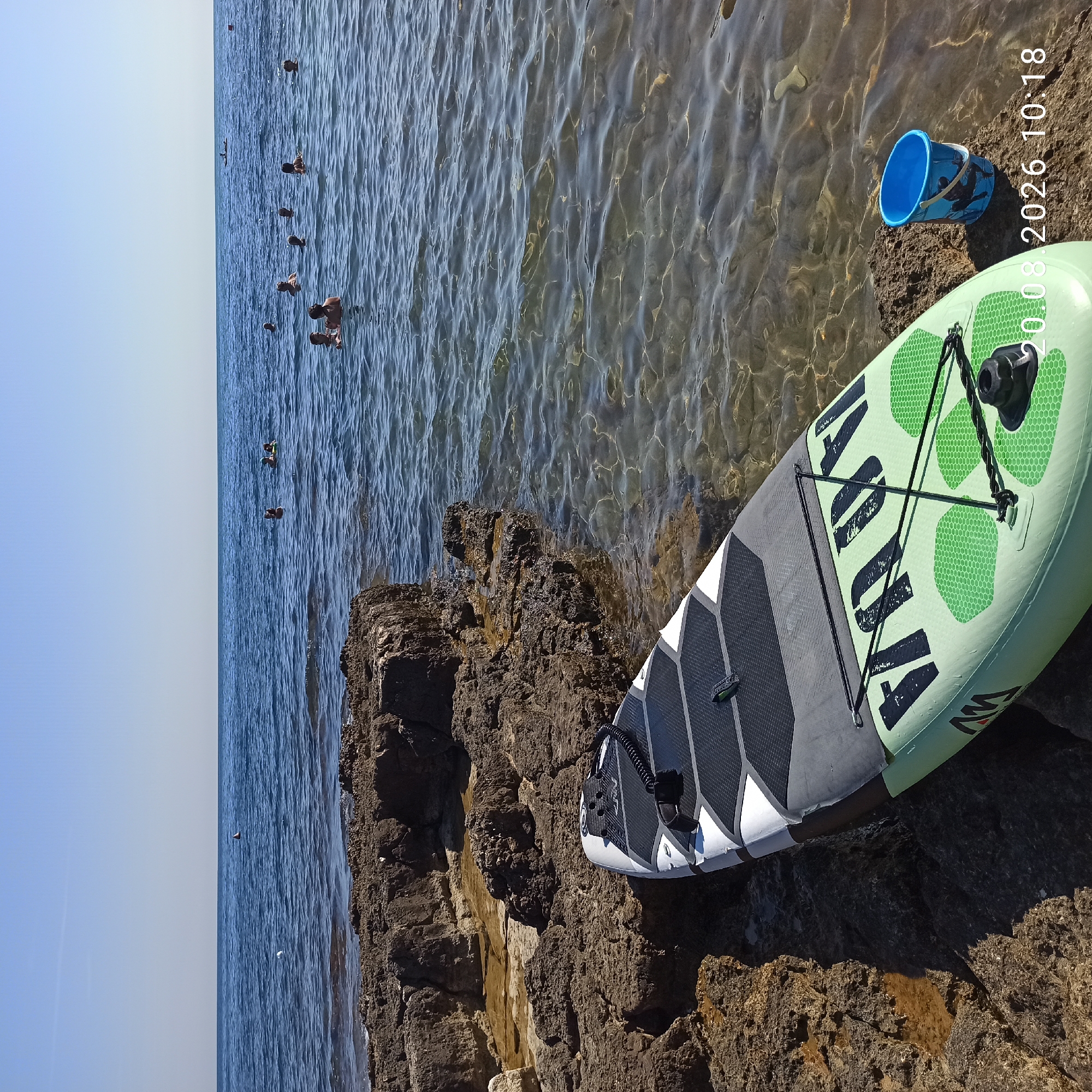 The image depicts a scene on the coast of Molfetta, Italy, on August 20, 2026, at 10:18 AM. In the foreground, a green and white stand-up paddleboard rests on a rocky shoreline. Beside the paddleboard, a blue bucket is placed on a rock. The sea is visible in the background with several people swimming or floating in the water. The sky is clear and blue. The environment is a coastal area with a rocky terrain leading to the sea.