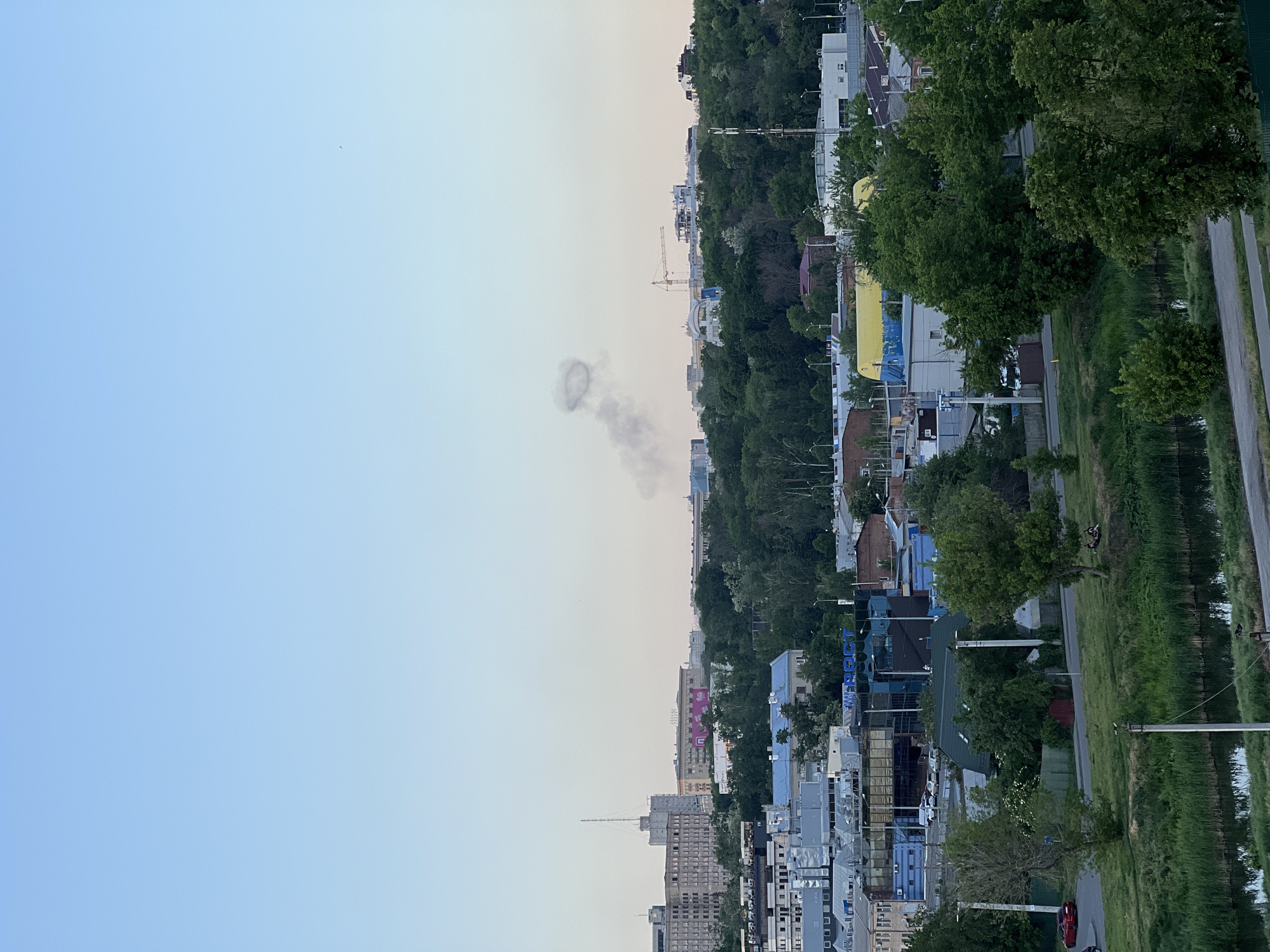The image depicts a city at dusk. In the foreground, a row of buildings varying in height and color transitions into a band of lush green trees along a river. A pathway runs alongside the river on the right side of the image. A smoke ring is visible in the sky slightly to the left of the center. There are also some structures that look like construction cranes in the distance.