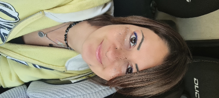 A close-up portrait depicts an adult female positioned inside a vehicle. The background features two dark grey or black headrests with white stitching, one partially displaying "DUCAT" in white capital letters, suggesting a Fiat Ducato or similar vehicle interior. The female has medium brown hair, brown eyes, and is looking directly at the camera with a subtle smile. She wears blue eyeliner on the upper lash line and cosmetic glitter is applied around her eyes and across the bridge of her nose. Visible facial piercings include a silver-toned horseshoe-style septum ring and a small silver-toned stud in her left nostril. She is dressed in a light yellow zippered hoodie, open to reveal a black garment underneath. She wears multiple necklaces: a black beaded choker, a silver-toned chain with a circular pendant, and another silver-toned chain with a small black pendant. No specific visual cues within the image indicate its location in the city of Tira, Israel.