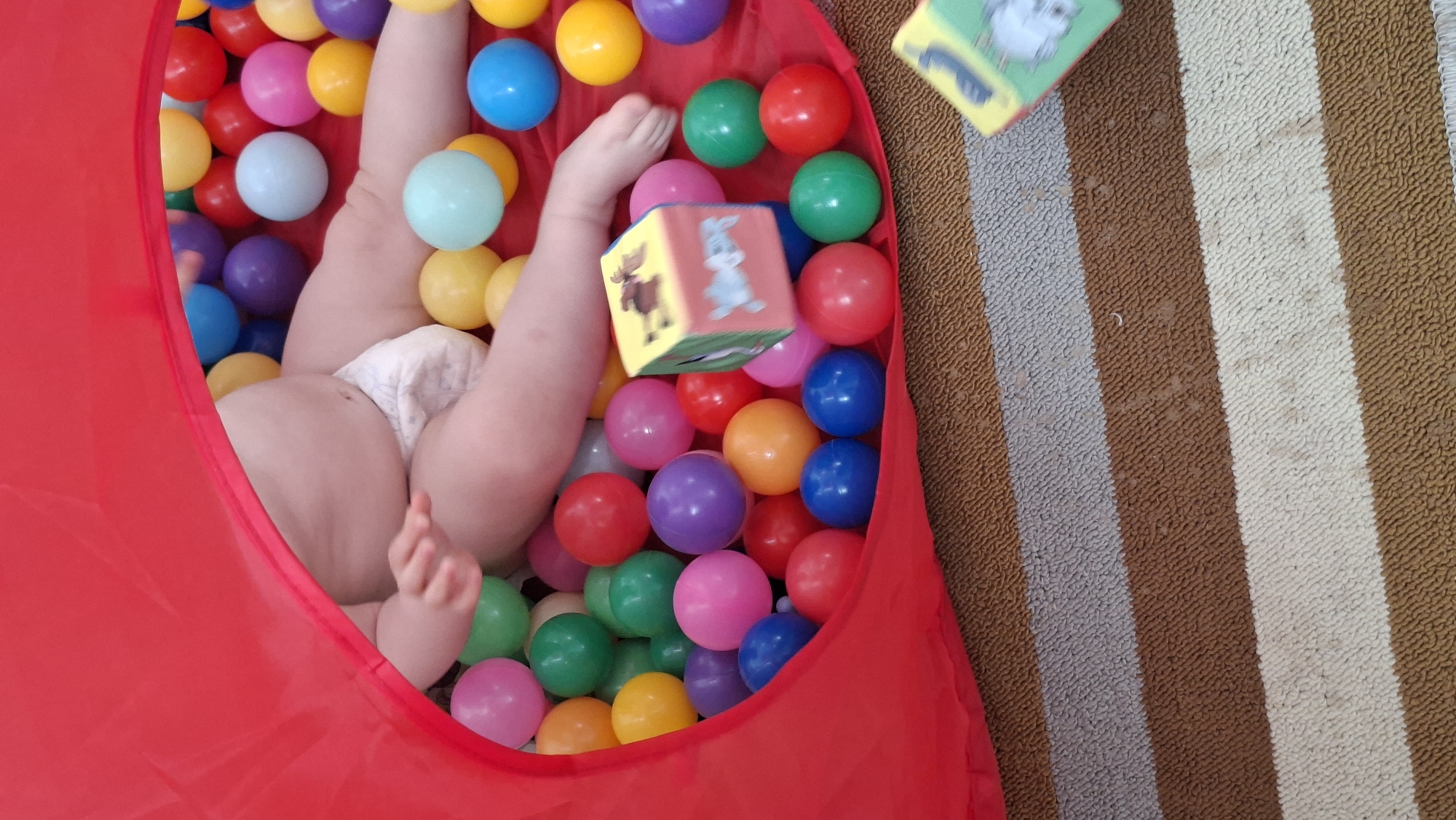 A baby wearing a diaper is partially submerged in a red ball pit filled with multi-colored plastic balls. Two toy blocks are in the air near the ball pit. The scene is situated on a striped rug of brown, white, and gray. The location of the scene is in the city of Chaikovsky, Russia.