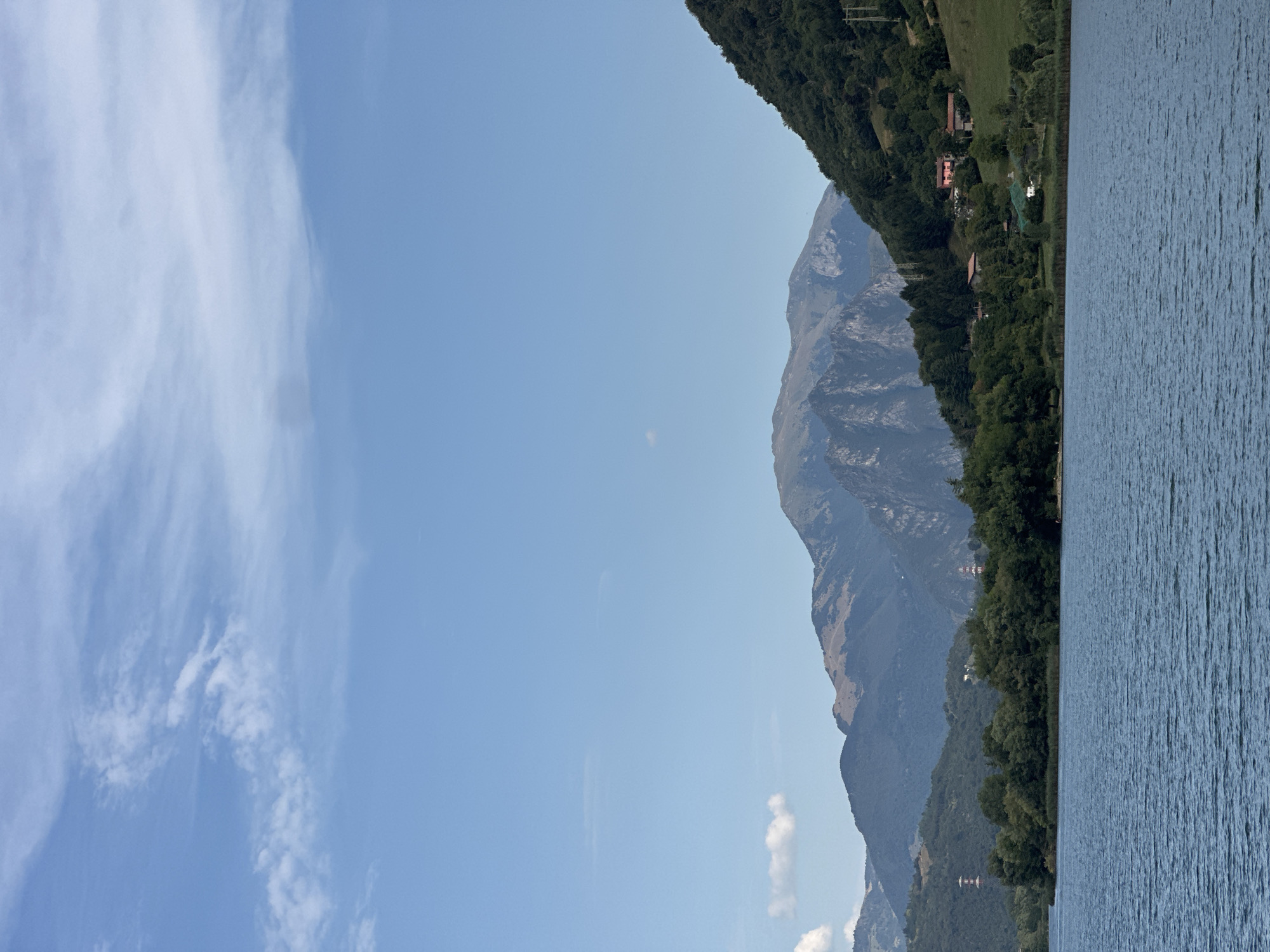This is a photograph of Lake Ranzanico, located in the municipality of Ranzanico, Italy. The image captures a wide shot of the lake's surface in the foreground, with gentle ripples reflecting the clear blue sky. In the middle ground, a dense line of trees borders the lake, with some taller trees interspersed. Beyond the trees, rolling green hills rise, dotted with scattered houses and buildings. The background is dominated by a series of majestic mountains, their peaks varying in texture and color, with some showing rocky outcrops. The sky is a bright blue, with scattered white clouds. The overall impression is one of a tranquil, natural landscape.