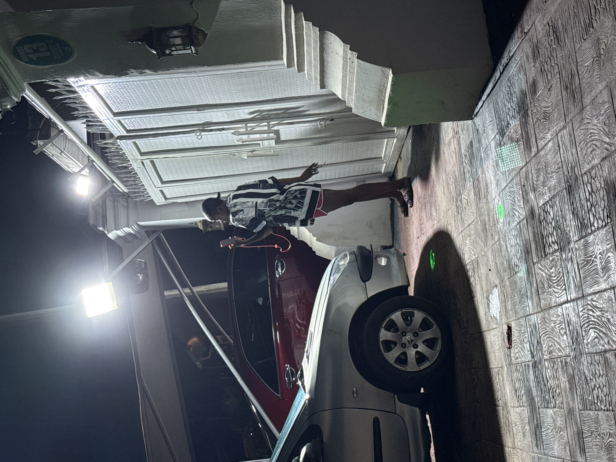 A nighttime outdoor scene in Abuja, Nigeria, features a woman standing near a white gate and a staircase. The woman is wearing a black and white patterned shirt and pink shorts. She is holding a pink mobile phone with a red cord. In the foreground, a silver car is parked. Behind it, a dark red car is visible. The ground is paved with patterned tiles, and there are lights illuminating the area. A sign on the wall reads "HOUSE 13 ZONE E." A decorative lantern hangs on the wall near the gate.
