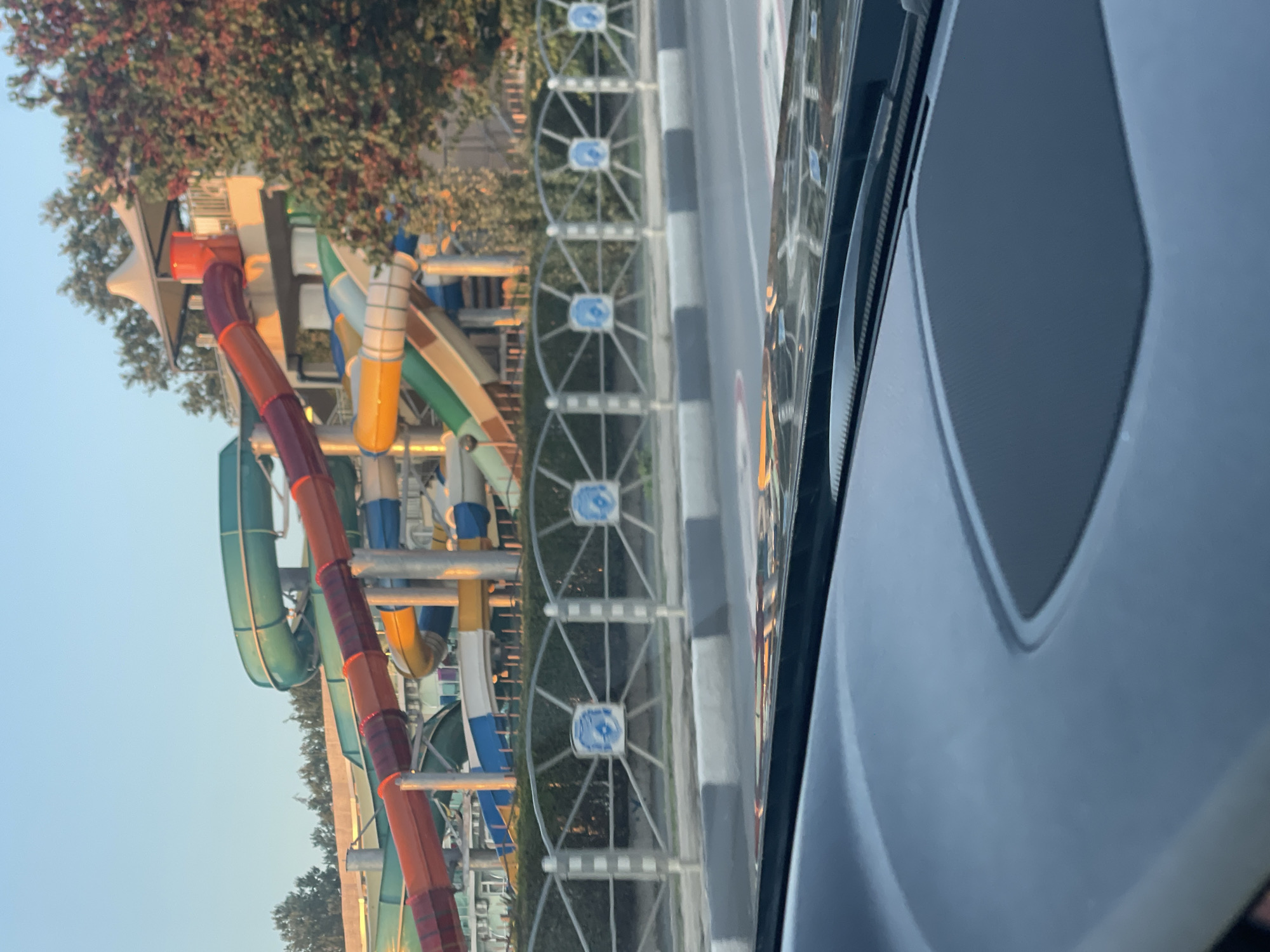 This image shows a view from a car's dashboard looking out at a water park in Nesebar, Bulgaria. The water park features several colorful water slides, including red, orange, yellow, green, and blue tubes, set against a backdrop of trees and a clear sky. A fence with circular emblems separates the water park from a road in the foreground. The car's windshield wipers are visible at the top edge of the frame. The lighting suggests it is either early morning or late afternoon.
