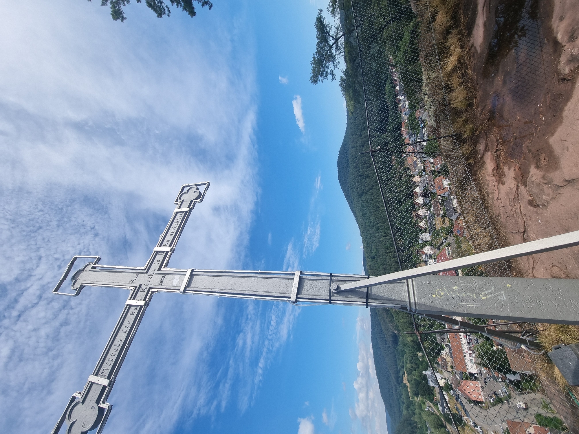 This image shows a tall metal cross positioned on a rocky outcrop overlooking the town of Dahn, Germany. The cross is inscribed with text, parts of which are legible and appear to read "Mitteln der K.-Kirche in L. errichtet 1872" which translates to "Erected by the middle of the K.-Church in L. 1872". In the background, a dense green forest covers a rolling hill, and the town of Dahn is visible below, featuring numerous houses with red-tiled roofs, a road with parked cars, and a small church spire. The sky above is a clear blue, dotted with scattered white clouds. A chain-link fence runs along the edge of the outcrop, separating the cross from the town and landscape below. The foreground is characterized by rough, reddish-brown rock formations and dry grass, with some puddles of water reflecting the sky.