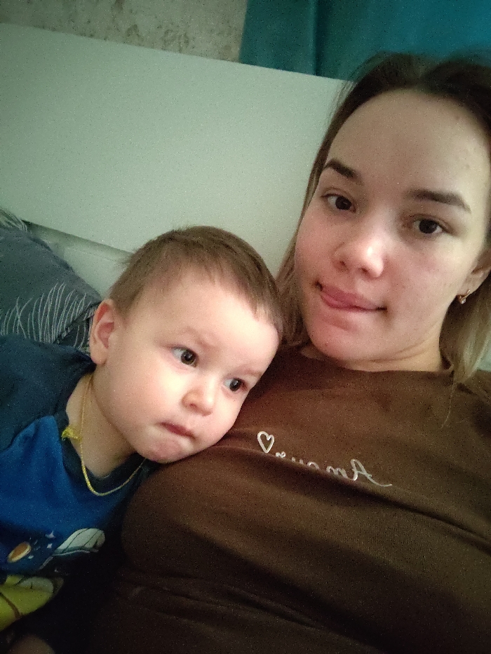 A woman takes a selfie with a young child. The child, wearing a blue shirt with yellow trim, rests his head on her chest. The woman wears a brown shirt with writing on it. She sticks her tongue out slightly, looking at the camera. They are both sitting, seemingly on a couch, with a pillow and white wall in the background. A blue curtain is slightly visible.