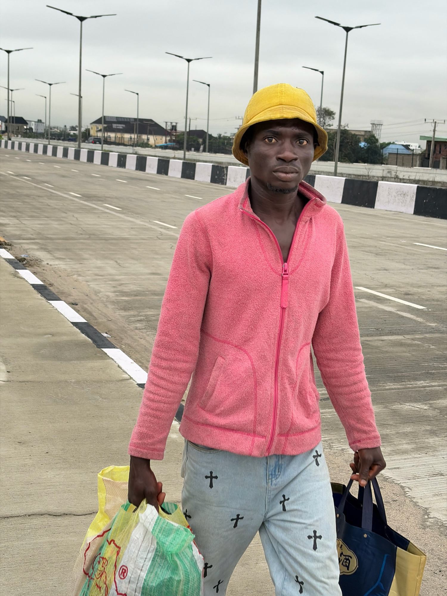 A young Black male wearing a yellow bucket hat, a pink fleece jacket zipped halfway, and light blue jeans with repeated black cross designs walks along a road shoulder in Lekki, Nigeria. He carries two bags. In his right hand, he holds a dark blue tote bag with some gold lettering. His left hand carries a large, multi-colored reusable shopping bag that is predominantly yellow, white, and green. The road is paved with asphalt and marked with white lines and a black and white striped barrier. Numerous streetlights with solar panels are visible in the background, along with buildings and trees. The sky is overcast.