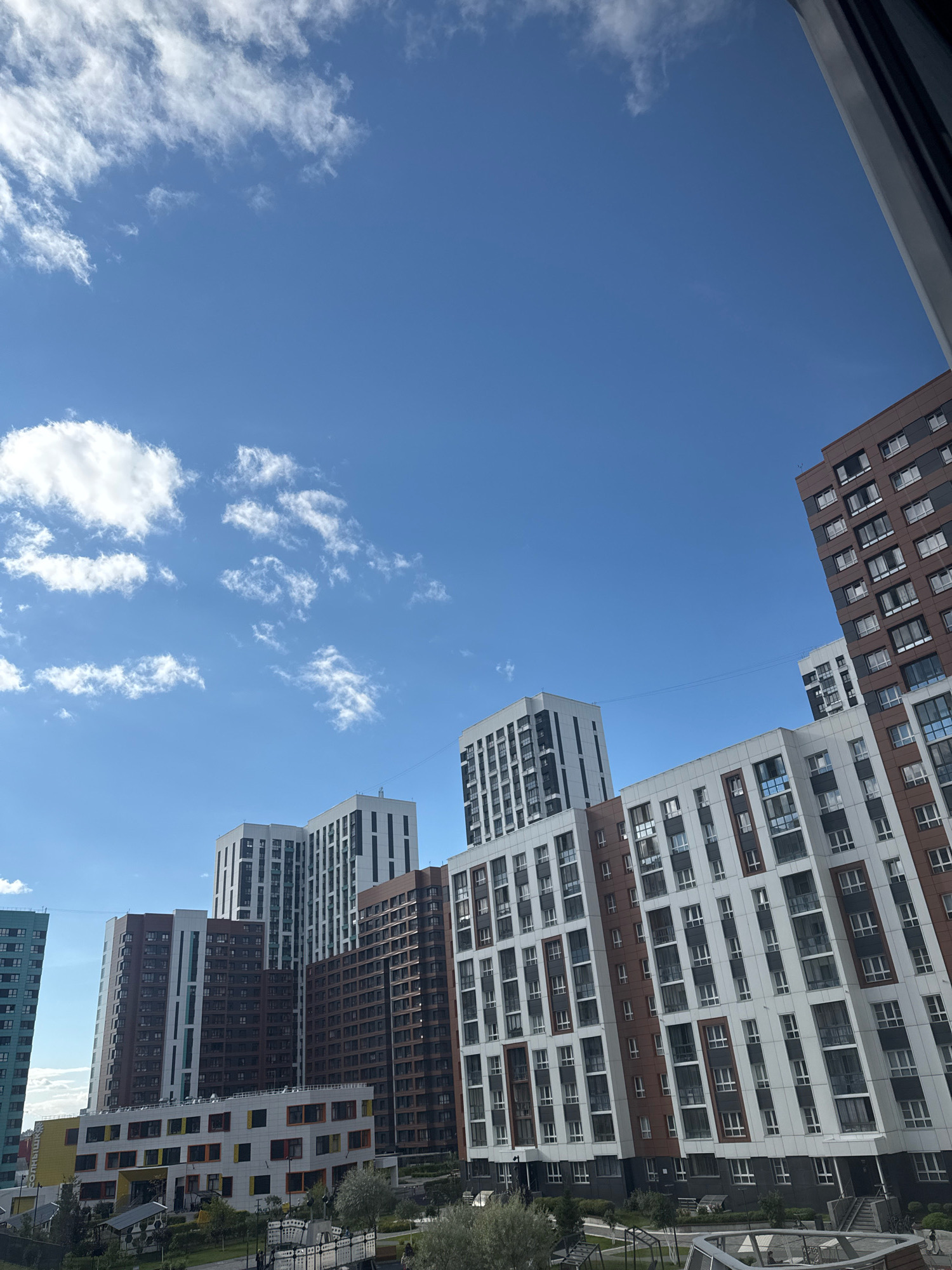 The image shows a bright, sunny day with a clear blue sky dotted with wispy white clouds. The scene is dominated by modern, multi-story residential buildings, some of which are quite tall and densely packed together. The architecture features a mix of materials, including brown, white, and grey facades, with numerous windows. In the foreground, a lower, colorful building with yellow and orange accents appears to be a school or kindergarten. Landscaped areas with trees and pathways are visible between the buildings. One building on the left has Cyrillic text on it, which reads "СОЛНЫШКО" (Solnyshko), meaning "little sun." This indicates the location is likely the "Solnyshko" residential complex in Novosibirsk, Russia. There are no discernible people or distinct actions taking place. The perspective is from a high vantage point, looking up at the buildings and the sky.