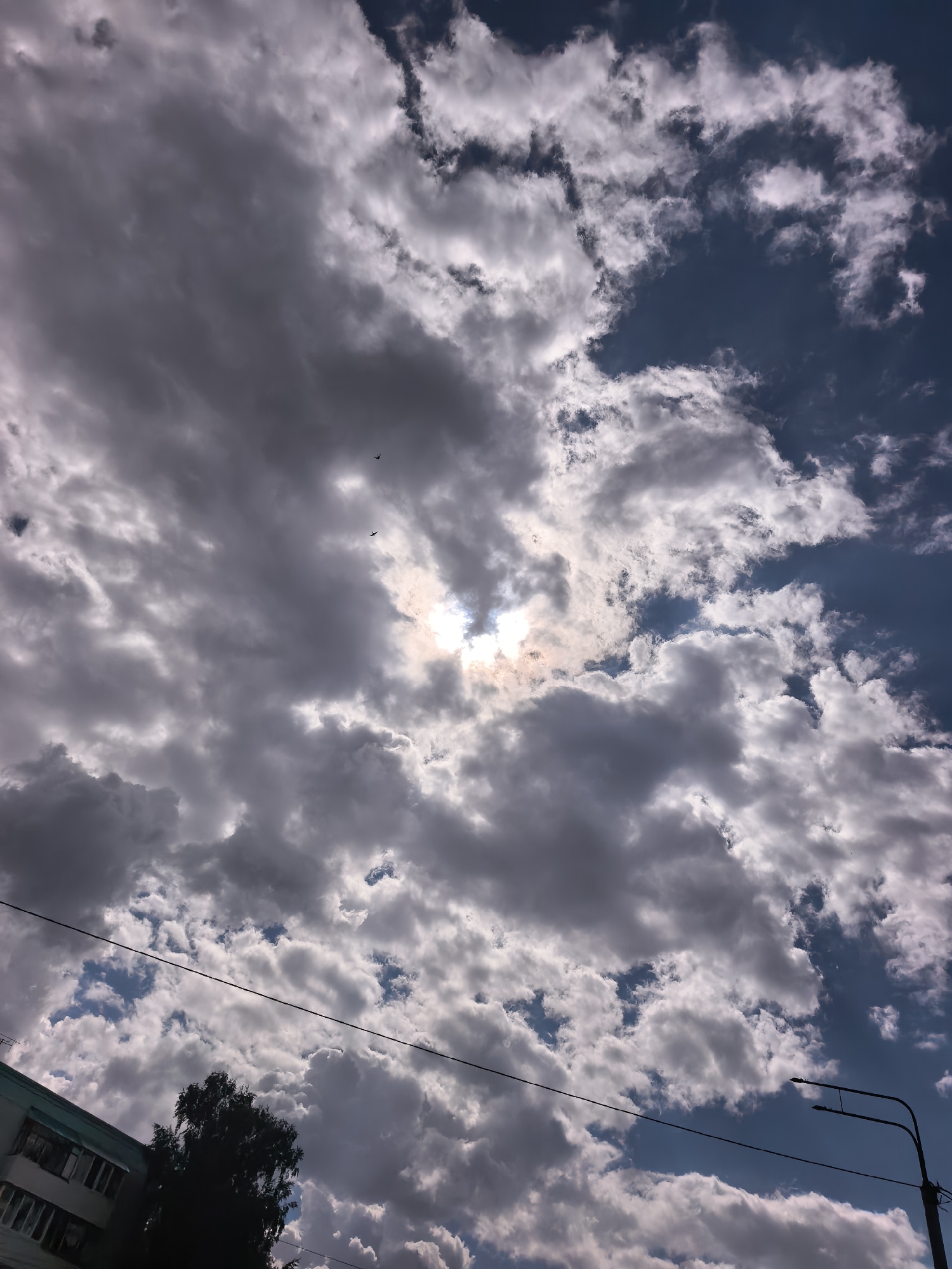 The image shows a cloudy sky with the sun partially obscured by clouds. In the lower left quadrant, a portion of a building with windows is visible, along with a dark tree. A power line stretches across the middle of the frame. In the lower right, a streetlight pole is present. Two birds are seen flying in the sky. The image is taken from a street-level perspective, suggesting an outdoor urban environment in Sterlitamak, Russia. The lighting indicates it is daytime.