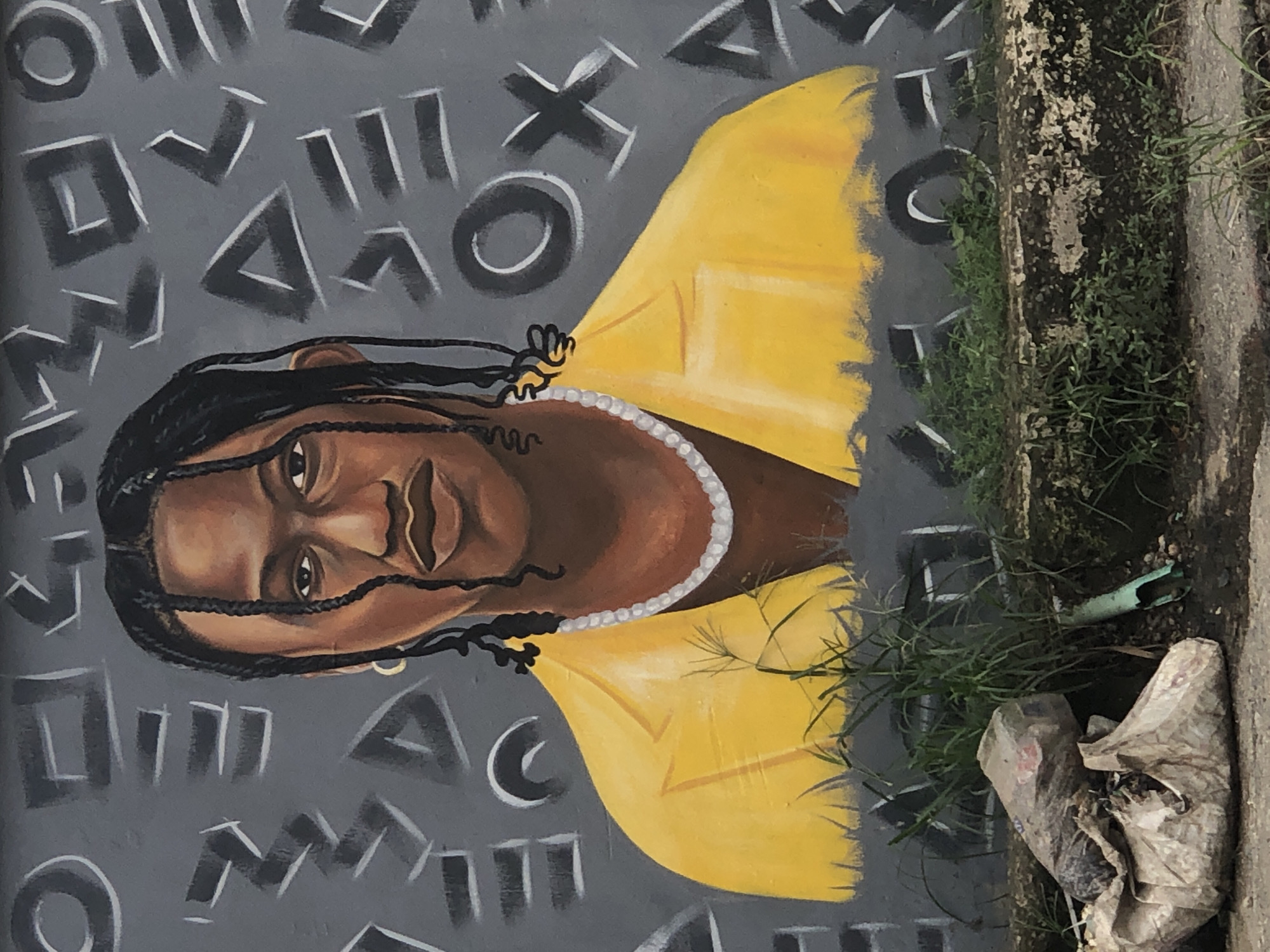A mural depicting a person with braided hair and a yellow jacket is spray-painted on a grey wall in Ikeja, Nigeria. The background of the mural features abstract shapes and symbols, resembling those found on video game controllers. The person in the mural is wearing a pearl necklace and gold earrings. In front of the wall, at ground level, is a pile of trash, including plastic bags and debris, with some green grass growing around it. The overall scene suggests an urban environment with signs of neglect and artistic expression.
