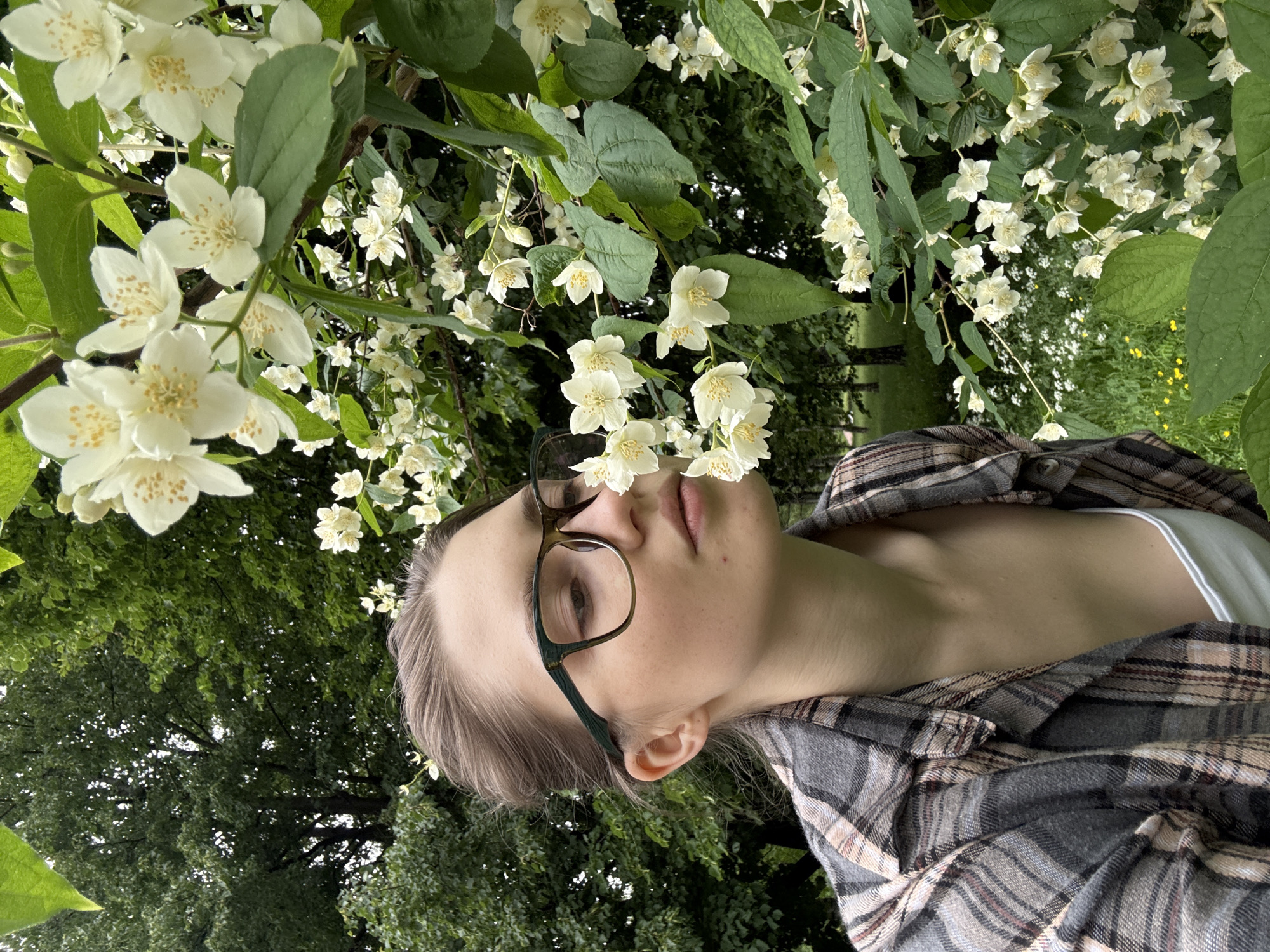 A young woman with brown hair pulled back, wearing glasses with dark green frames and a plaid shirt over a white tank top, is shown from the chest up. She is standing outdoors, with her head turned to her right and her nose close to a branch of white jasmine flowers. The background is a green, leafy environment, suggesting a park or garden setting. The image does not provide any specific clues about the location within the city of Saint Petersburg, Russia.