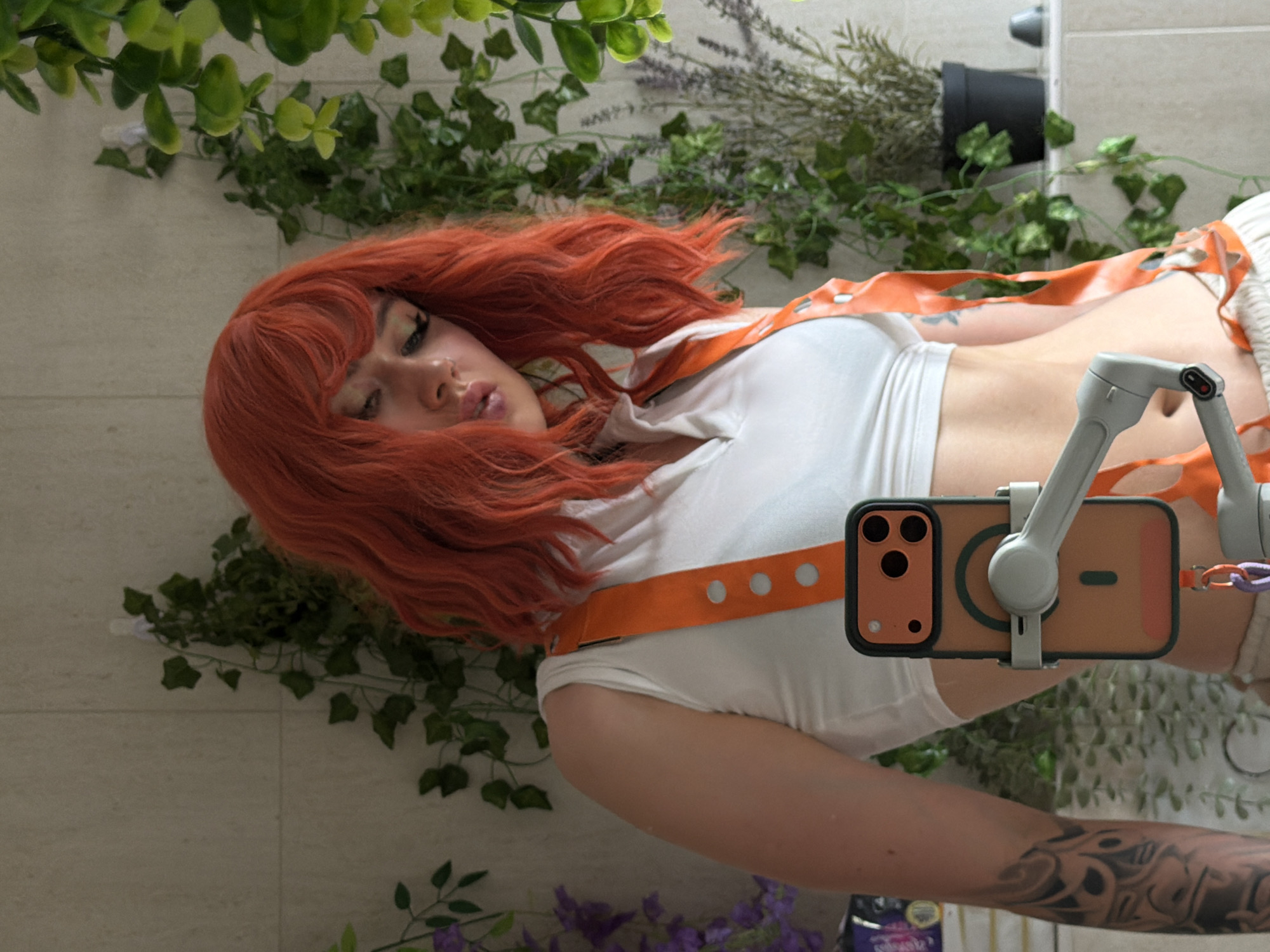The image, taken in Budva, Montenegro, captures a person with vibrant, wavy red hair, positioned horizontally across the frame. The individual wears a white crop top and an eye-catching orange harness or straps featuring circular cutouts, which span across their torso. Their head is oriented to the left, with eyes mostly closed and lips slightly parted, conveying a serene and stylized expression. A detailed dark-patterned tattoo is visible on their left forearm.
The person is actively holding a light-colored gimbal or stabilizer, which firmly secures an iPhone. The smartphone, encased in an orange cover with a black border, displays its triple camera system prominently. The background consists of a light-toned tiled wall, artfully decorated with strands of artificial green ivy-like foliage and small clusters of purple flowers, resembling lavender. A black pot containing additional green plants is visible in the upper right. The overall scene suggests an indoor setting, possibly a bathroom or a decorated room, with diffused indoor lighting. A small label or package in the bottom right corner has partially visible text including "pure" and "beauty." The composition implies a posed moment, possibly a selfie or video capture.