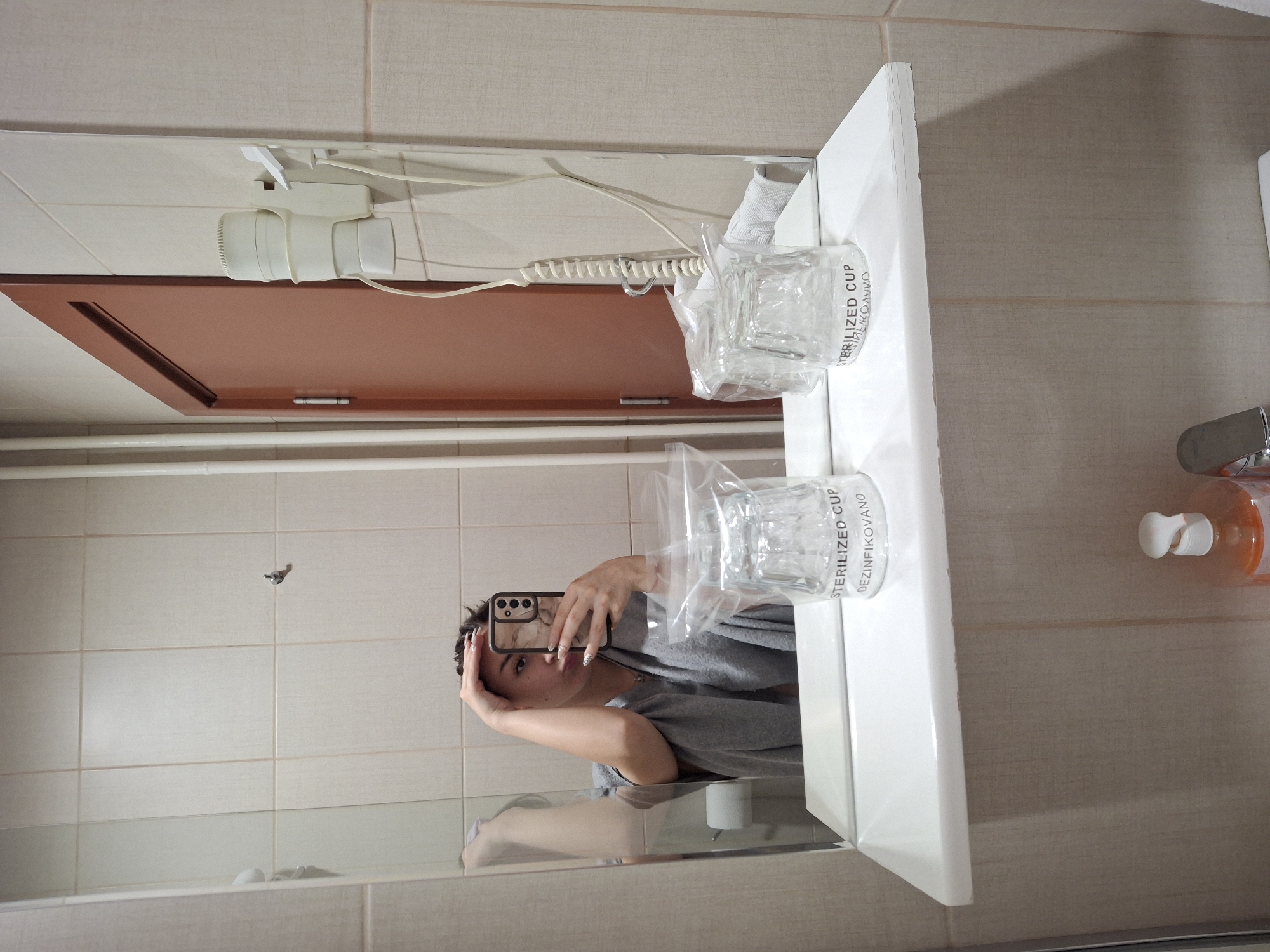 The image displays a mirror selfie taken within a bathroom, likely situated in a hotel or accommodation in Vozdovac Urban Municipality, Serbia. A single individual, appearing female, is reflected in a rectangular wall-mounted mirror. She has short, dark hair and wears a grey garment. Her right hand (left in reflection) holds a smartphone with a light-colored, patterned case, partially obscuring her face as she takes the photograph. Her left hand (right in reflection) rests near her forehead. Her fingernails are manicured with light-colored polish and dark accents.
A white, rectangular shelf is mounted directly below the mirror. On this shelf, two clear glass cups are present, each individually sealed within transparent plastic bags. The bags are labeled "STERILIZED CUP" and "DEZINFIKOVANO" (disinfected in Serbian).
The background elements visible in the mirror reflection include walls clad in light-colored, rectangular tiles. To the left of the reflection, a light brown/reddish door with a visible hinge is present. To the right, a white wall-mounted hairdryer with a coiled power cord is affixed to the tiled wall. A white hand towel is partially visible draped beneath the hairdryer. In the far-left portion of the reflection, a toilet paper dispenser with a roll of toilet paper is partially visible. Below the white shelf, parts of a chrome faucet and a bottle containing an orange liquid with a white pump dispenser are visible.