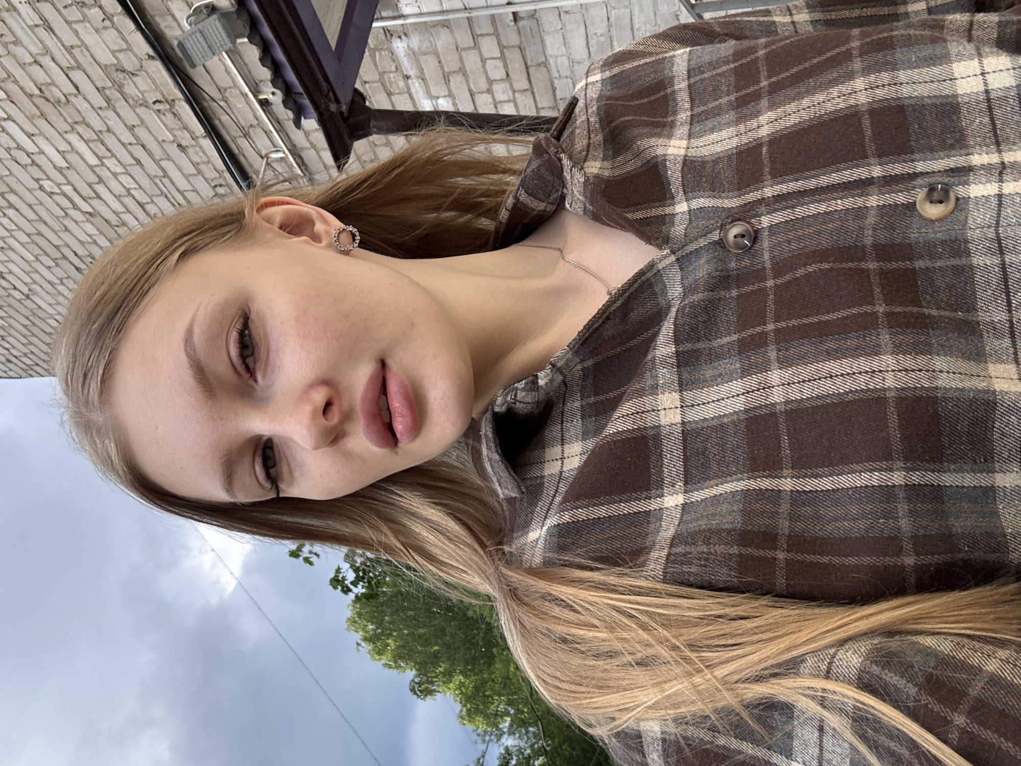 A young woman with long blonde hair is posing for a selfie. She is wearing a brown and gray plaid shirt. She has a silver hoop earring in her left ear and a delicate silver necklace. Her skin is fair and she has neutral-colored lipstick. The background includes a brick wall, a dark-colored overhang with metal elements, trees, and a cloudy sky. The location is Perm, Russia.