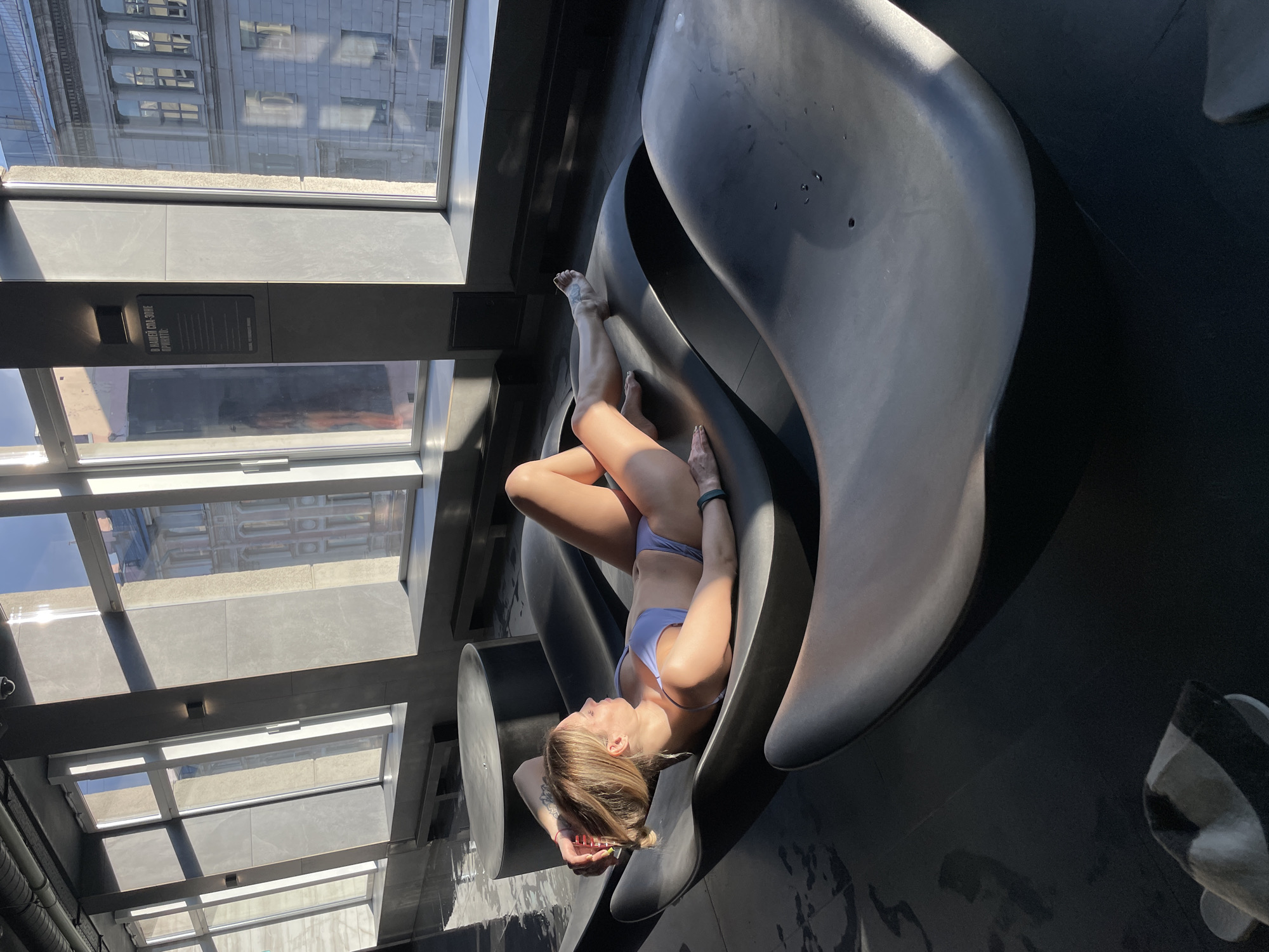 The image depicts a woman in a light purple bikini lounging on a curved, black chaise lounge in an indoor swimming pool or spa area. She is lying on her back with her head propped on her hand and her hair in a bun. The chaise lounge is positioned near a large window that overlooks a city street with ornate buildings, consistent with the architecture found in Saint Petersburg, Russia. Sunlight streams through the window, casting shadows and highlighting the water on the floor. There are other similar chaise lounges visible, as well as some dark circular furniture in the background. A sign with Russian text is affixed to the wall near the window. The overall atmosphere is one of relaxation and tranquility.