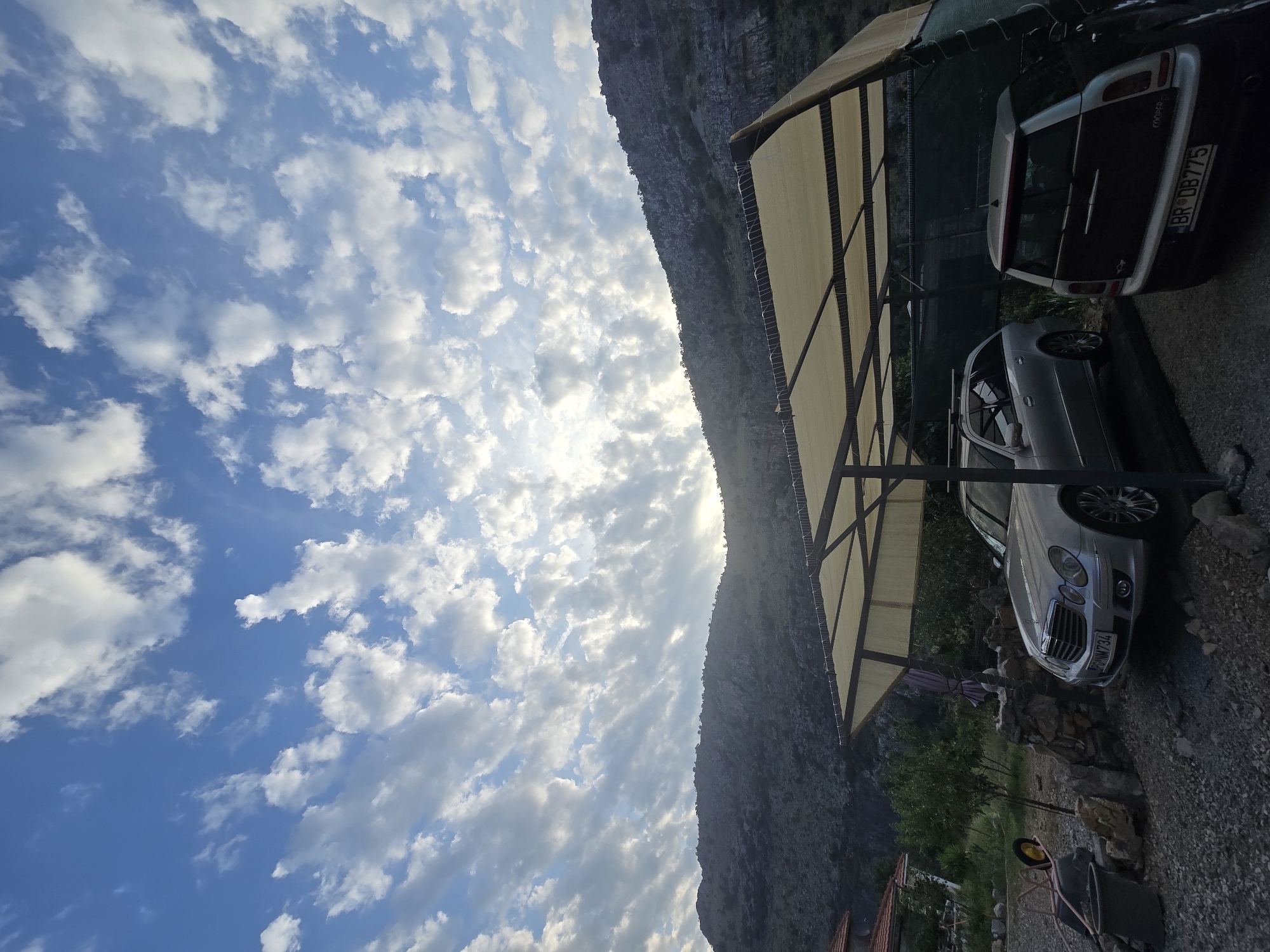 The image captures an outdoor scene in Podgorica, Montenegro, under a cloudy sky with sunlight peeking through. The foreground features a gravel driveway with two parked cars under a beige canopy supported by dark metal poles. A silver Mercedes-Benz station wagon is parked on the left, and a dark Mini Cooper is on the right, its rear visible. The cars have license plates. To the left of the canopy, there is a stone wall, a wheelbarrow with a yellow wheel, and a gray bin. In the background, a lush green hillside with trees rises behind the cars, and further back, a rocky mountain range dominates the horizon. The overall setting suggests a residential or semi-rural area, possibly a driveway or courtyard attached to a property. The time of day appears to be either early morning or late afternoon, given the lighting conditions. There are no people visible in the image.