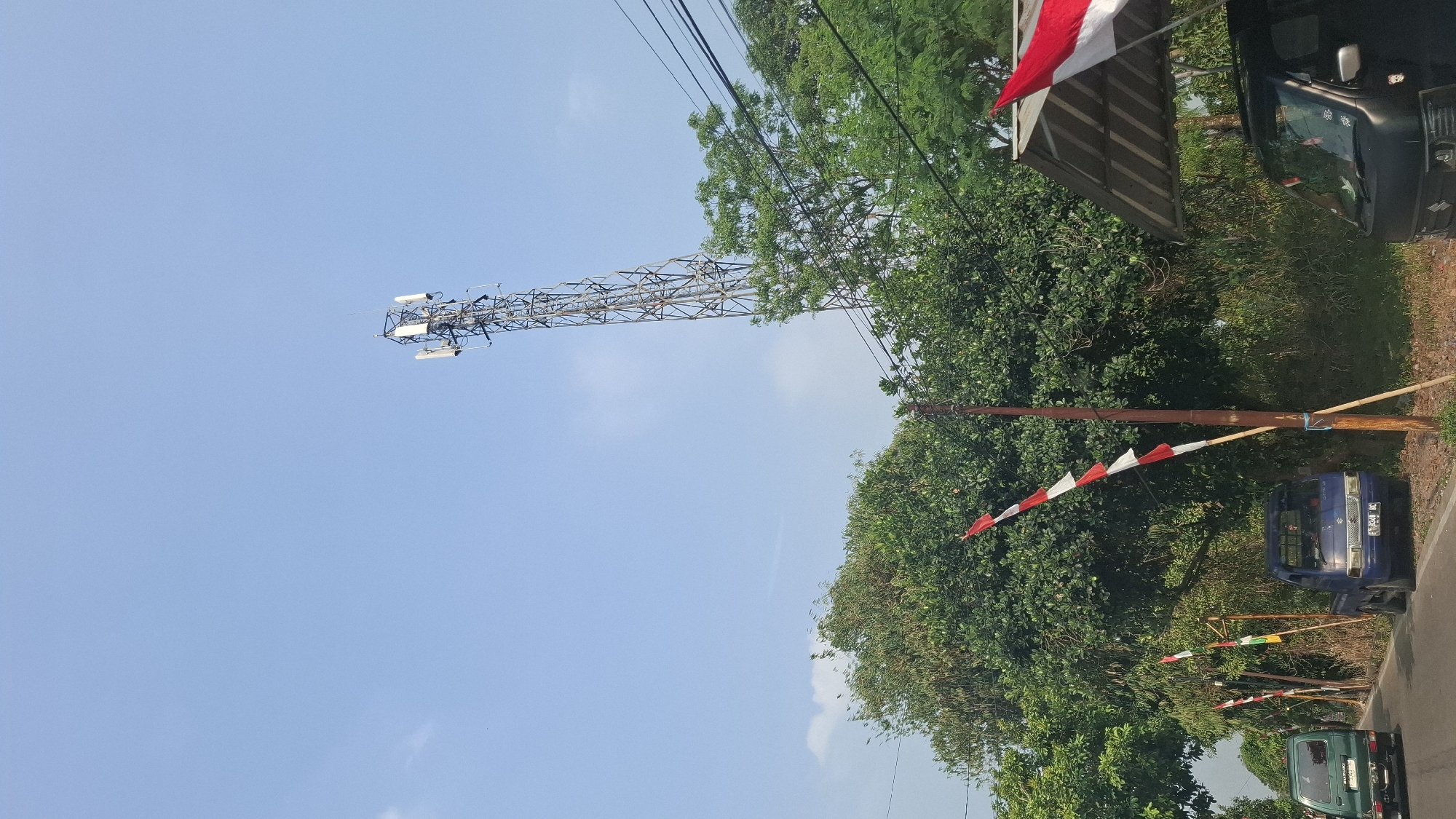 The image shows a clear blue sky with a tall telecommunications tower in the background. In the foreground, there are lush green trees and a road. A blue pickup truck is parked on the side of the road, and a green vehicle is driving away. Indonesian flags, red and white, are displayed on poles along the side of the road, suggesting a festive or nationalistic occasion. A small shelter with a corrugated roof is also visible. The scene appears to be in Cibinong Hilir, Indonesia, based on contextual information not present in the image itself. There are no people clearly visible in a way that would indicate specific actions or interactions. The environment is rural or semi-rural, with a focus on infrastructure and natural elements.