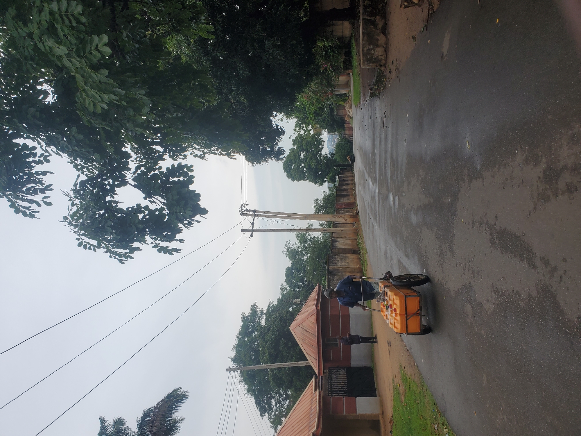 In Jos, Nigeria, on a wet asphalt road under an overcast sky, a man pushes a trolley laden with several yellow jerrycans. He is dressed in a blue short-sleeved shirt and light-colored shorts, wearing a head covering. To his left, a person stands near a gate entrance to a building with a red, sloped roof, dressed in dark clothing. The street is bordered by walls and lush green trees, with power lines crisscrossing the sky.
