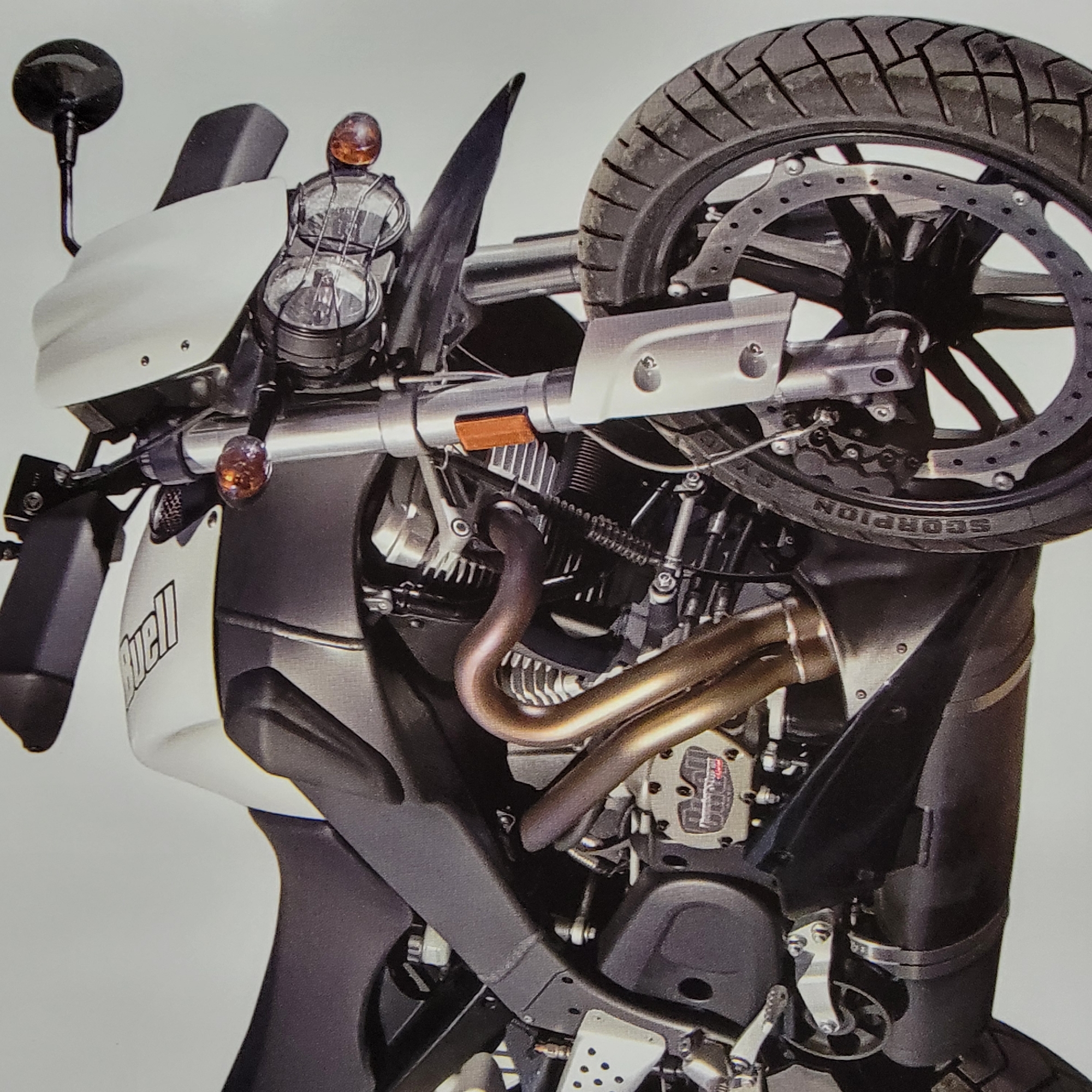 The image is a close-up, overhead shot of a motorcycle, with the bike angled and partially cropped. The motorcycle is mostly dark gray and black, with some white fairings and accents. The prominent engine components are visible, including copper-colored exhaust pipes and intricate mechanical parts. The rear tire, with the brand name "SCORPION" visible on its sidewall, is a focal point, with its tread pattern clearly discernible. The motorcycle's headlight assembly and a side mirror are also visible towards the top of the image. The overall aesthetic suggests a focus on the engineering and design of the motorcycle. There is no visible indication of people, an event, or specific environmental conditions like time of day or weather. The background is a plain, light gray, which isolates the motorcycle as the sole subject. Visible text includes the brand name "Buell" on the white fairing and "SCORPION" on the tire.