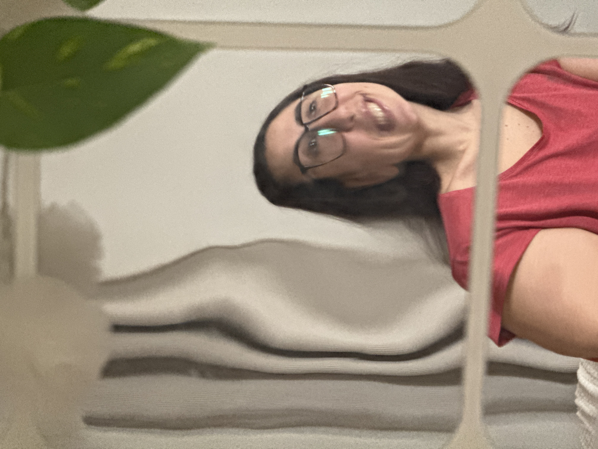A woman with dark hair and glasses, wearing a red shirt, is visible through a partially obscured frame. She is smiling. A green leaf is present in the foreground, obscuring part of the scene. In the background, there is a soft, grey, undulating surface that appears to be fabric. The image does not provide enough information to identify the city of Krasnoyarsk, Russia.