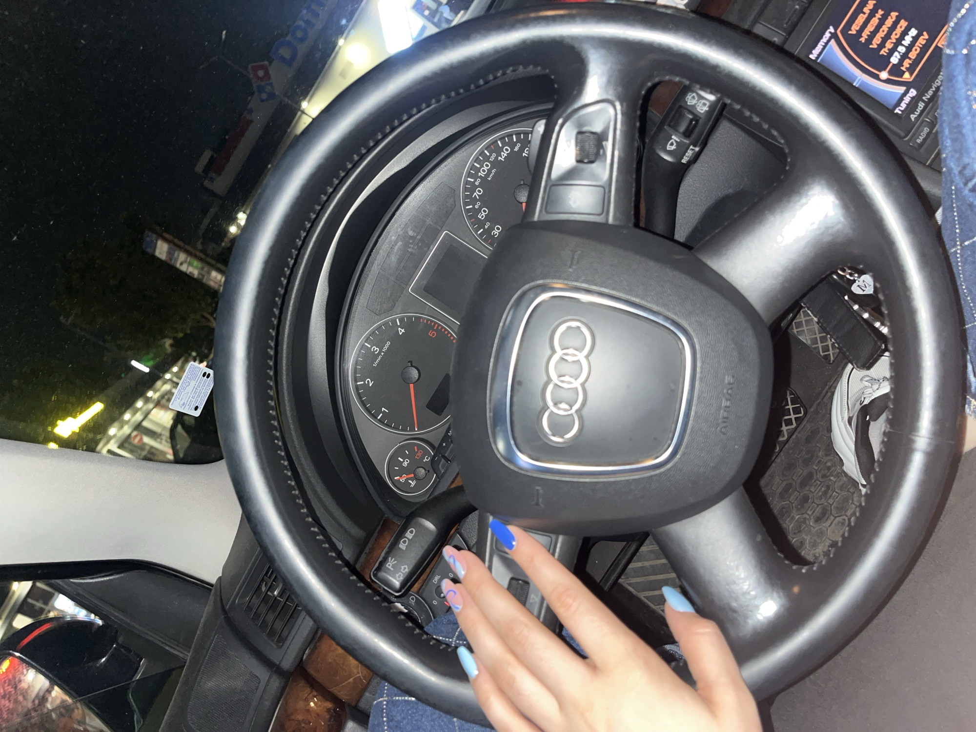 The image displays the interior of an Audi vehicle, primarily from the driver's perspective, at night in a stationary position. A person's left hand, with nails painted in varying shades of blue, rests on the black leather-wrapped steering wheel, which prominently features the Audi four-ring emblem. The dashboard includes a speedometer displaying up to 260 km/h, a tachometer, and a temperature gauge indicating an operating temperature of approximately 90°C. A turn signal/light stalk is visible on the left, set to "DRL," and a wiper control stalk is on the right. The dashboard features dark wood-grain trim. Below the steering wheel, a dark, patterned floor mat is visible, along with a black keychain bearing a silver "M" charm. The car's infotainment system, labeled "Audi Navigation," displays radio information, including current and saved stations such as "VESELINA," "FRESH," "VERONIKA," "THE VOICE," "87.5 MHz," and "HR.BOTEV."
Through the windshield, a nocturnal urban environment is visible, illuminated by numerous artificial lights. A large, brightly lit "Domino's Pizza" sign, displaying its characteristic red, white, and blue logo, is clearly identifiable in the background. Other indistinct commercial signs and building lights are also present. Based on the visible Bulgarian radio stations on the infotainment system and the presence of a Domino's Pizza, the scene is located in a commercial area within the city of Plovdiv, Bulgaria, in the immediate vicinity of a Domino's Pizza restaurant.