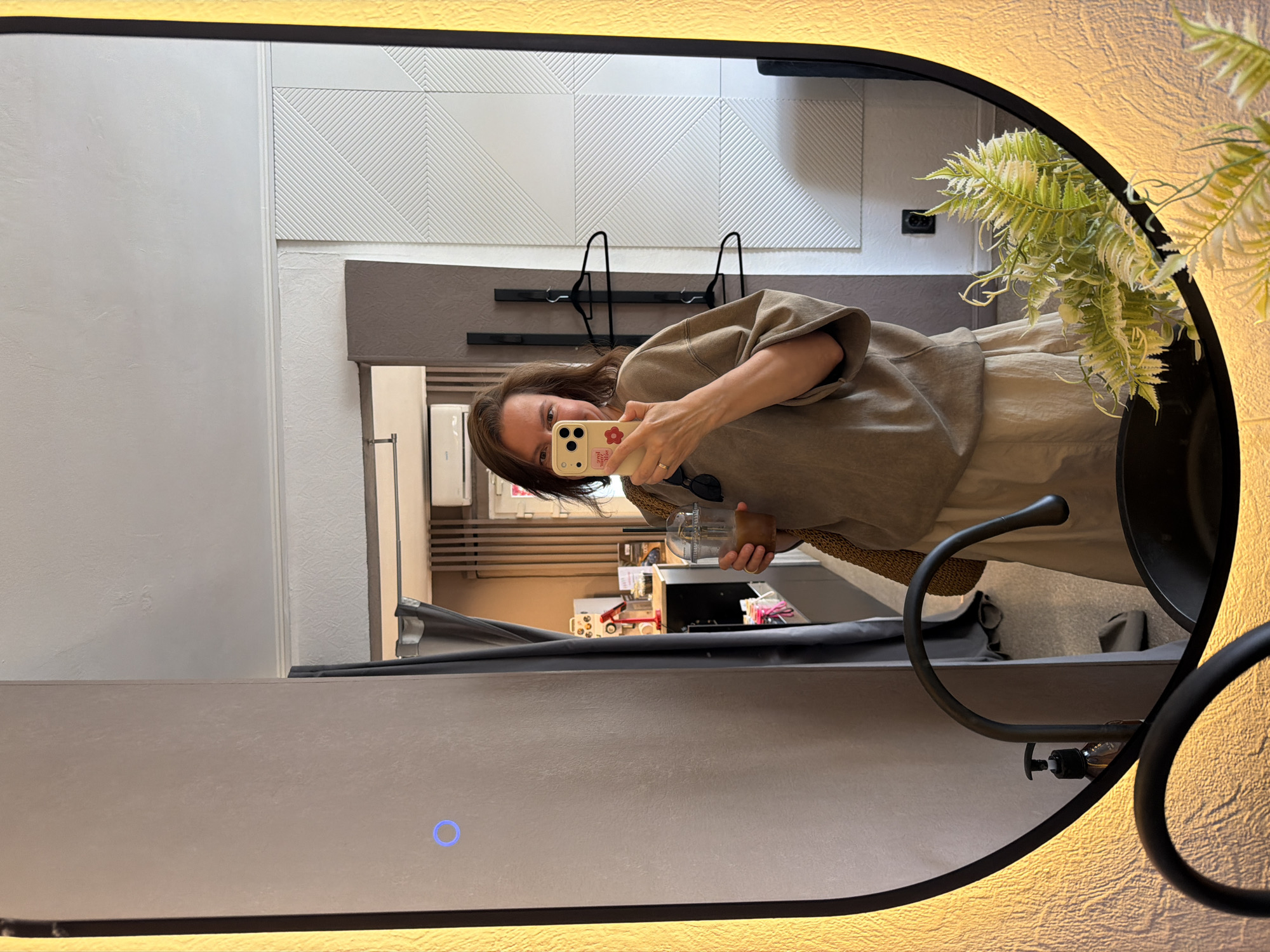 The image captures a woman taking a mirror selfie in what appears to be a hotel room or a similar accommodation. The woman is holding a phone in her right hand, taking the picture, and a cup with a straw in her left hand. She is wearing a loose-fitting, light brown top and a beige skirt. The mirror is large, oval-shaped, and illuminated by a warm LED light around its frame. The background reflected in the mirror shows a room with white textured walls, a white patterned wall panel, a curtain, and some hangers on the wall. A sink and faucet are visible in the foreground of the mirror's reflection. A plant with green and white fern-like leaves is placed in the lower right corner of the mirror, also reflected. The overall impression is a well-lit, modern, and tidy space. No specific identifiable landmarks or features suggest the location is Chaikovsky, Russia.