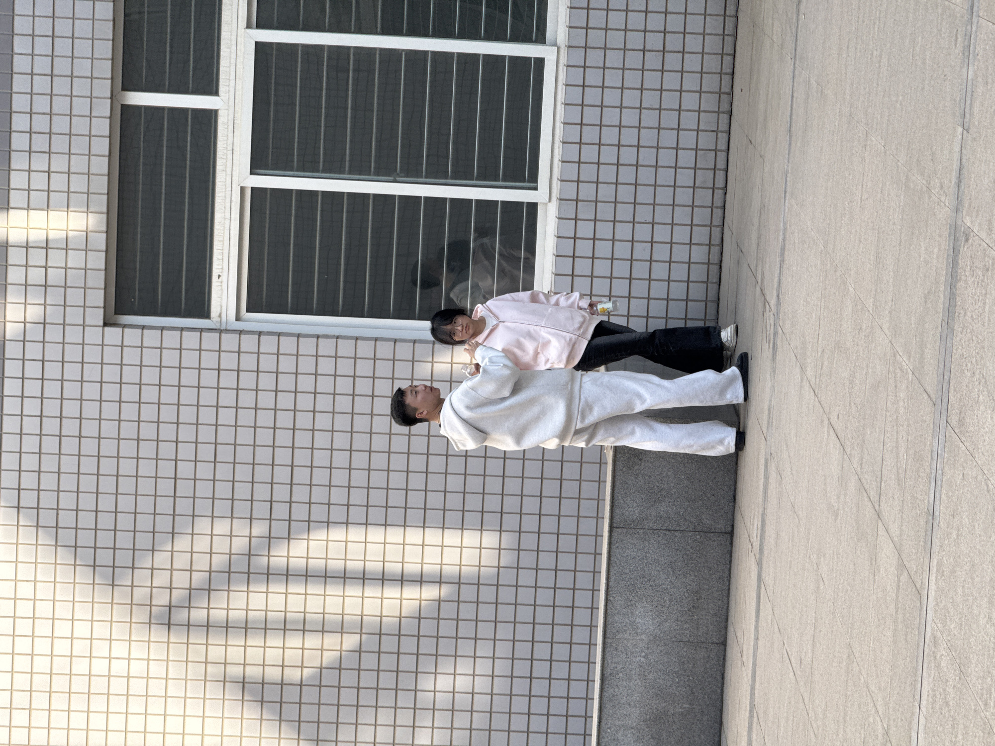 The image shows two people, a younger individual and an older one. The younger person wears a pink jacket and black pants and appears to be holding a beverage. The older person is wearing a gray tracksuit. The background consists of a building with a barred window and tiled exterior, as well as a paved area. The location is inferred to be Zhongtun, China.