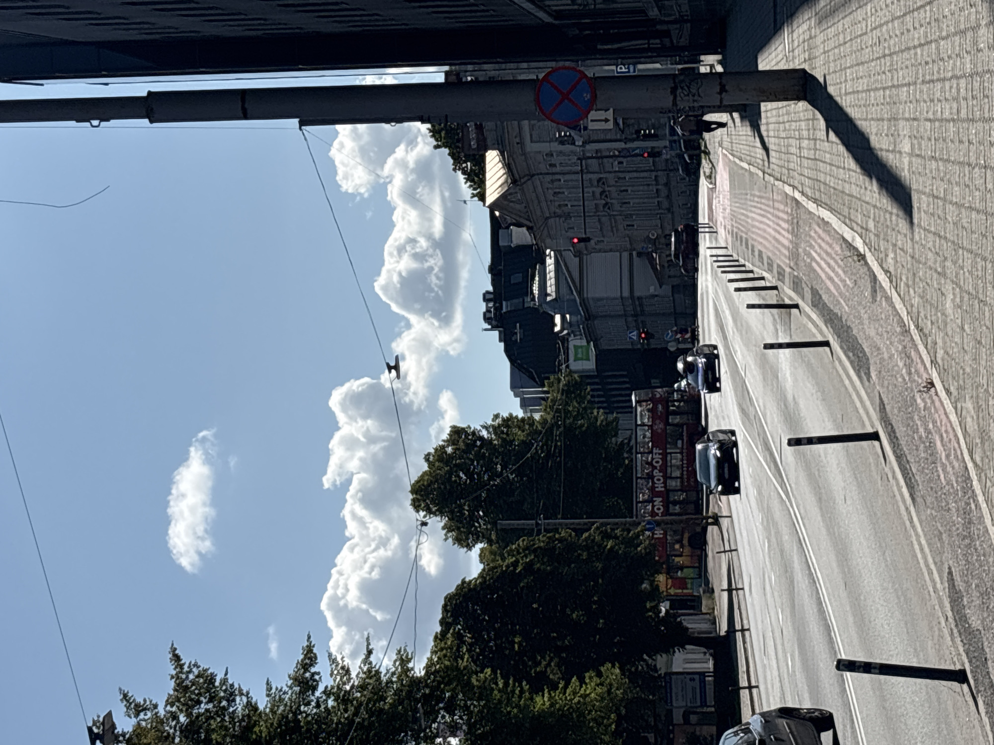 This is a street scene in Tallinn, Estonia, on a sunny day with a blue sky dotted with white, puffy clouds. A multi-lane road runs diagonally from the lower left to the upper right of the frame. Several cars are on the road, including a dark-colored sedan in the middle lane, and another dark-colored car further ahead. A red double-decker tour bus with "HOP-ON HOP-OFF" visible on its side is parked or moving slowly on the left side of the road.
To the right of the road is a paved sidewalk with bollards marking its edge. A person, visible from the back, is walking along the sidewalk away from the camera. A lamppost casts a long shadow across the sidewalk. A traffic sign with a red circle and a blue cross inside, indicating no stopping or parking, is mounted on a pole on the right side of the sidewalk.
In the background, there are several buildings. On the left, behind the tour bus, are trees and some buildings. Towards the center and right, there are multi-story buildings with architectural details, some appearing to be older stone structures, while others are more modern. Traffic lights are visible, showing red. Overhead, power lines stretch across the sky, supported by poles. The overall impression is a busy urban environment on a clear day.