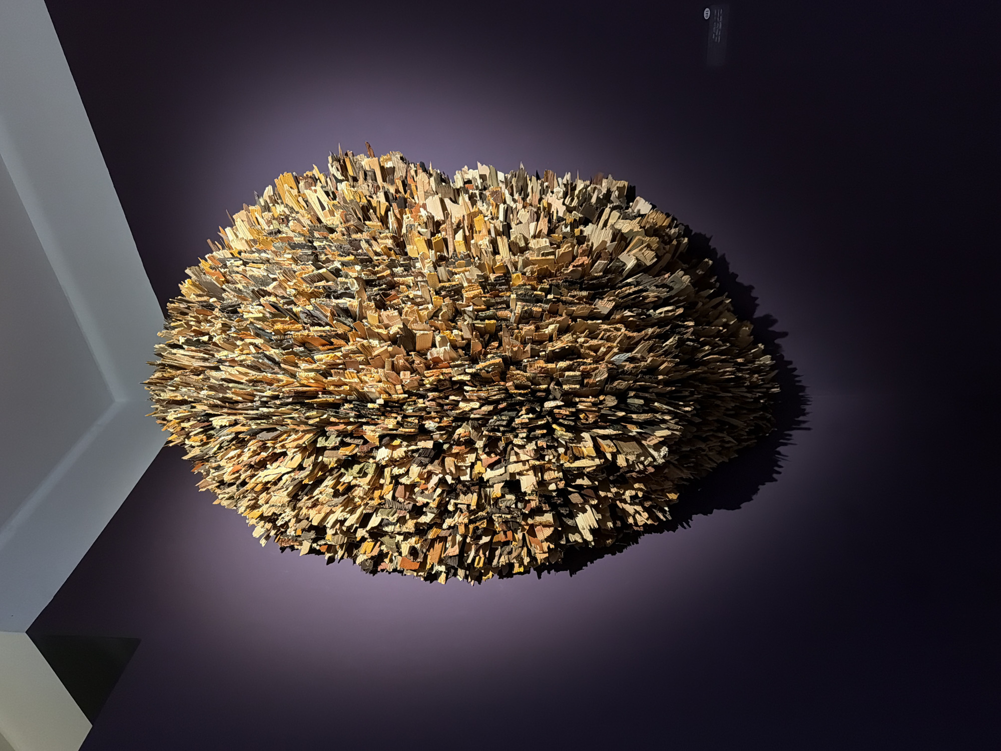 An eye-level shot depicts a spherical art installation on a dark violet wall. The installation is made of numerous small, rectangular pieces of wood or similar material in varying shades of brown, tan, and black. The pieces are arranged densely, creating a textured, somewhat spiky appearance. To the left of the art installation is a white wall and angled ceiling. The shadow of the installation is cast on the violet wall to the right. The location is presumed to be in Tel-Aviv, Israel.