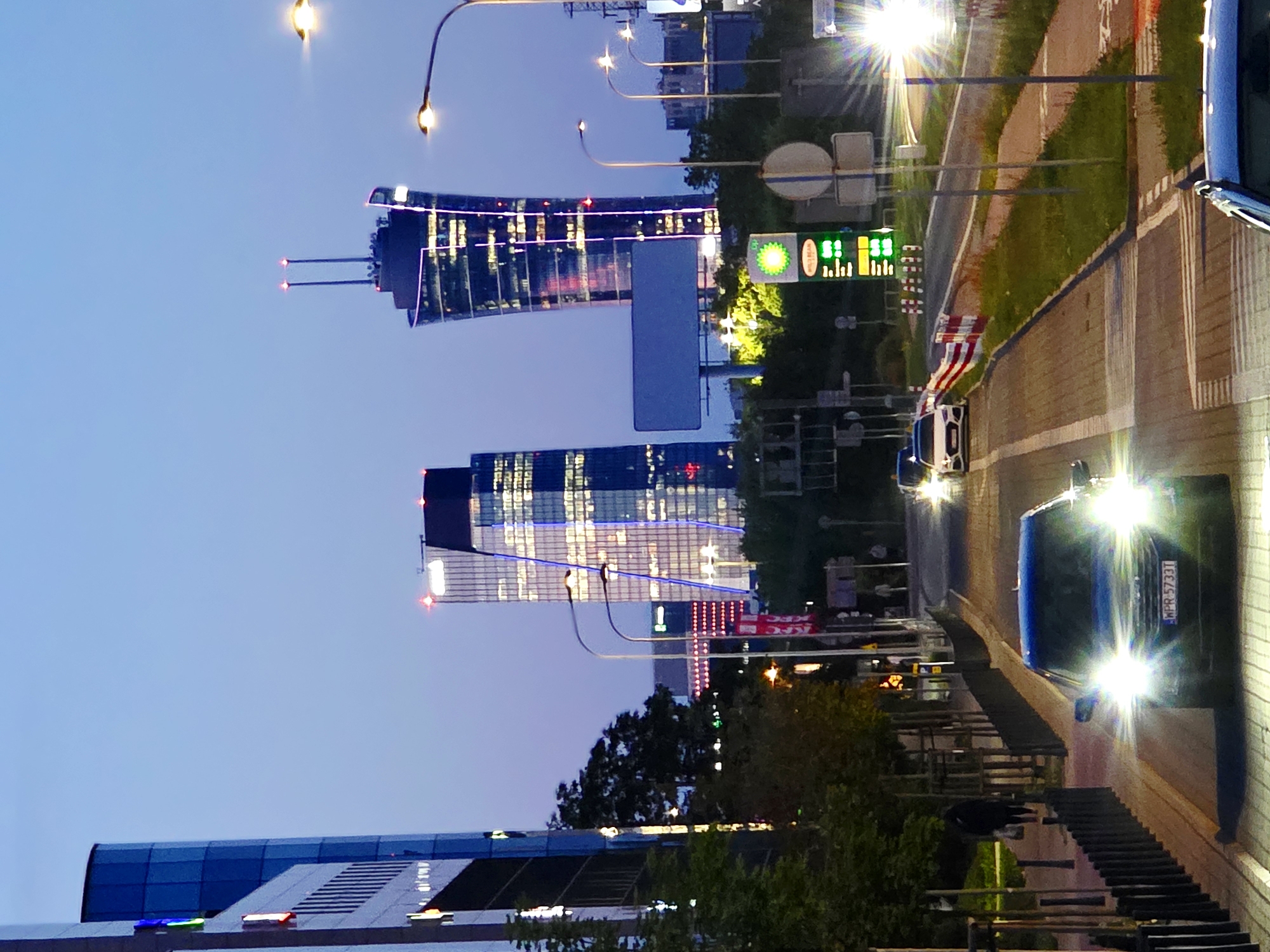The image depicts a street scene at dusk in an urban environment. In the foreground, a blue car with its headlights on is driving towards the viewer, its license plate reading "WPR 5733T". To the left, a sidewalk with trees and a black metal fence lines the street. On the right, there are more streetlights, a gas station sign with the BP logo and prices, and road construction barriers. In the background, several modern skyscrapers are visible, lit up and reflecting the twilight sky. The sky is a deep blue, transitioning to a lighter hue near the horizon. The overall impression is of a city street at the beginning of the evening, with traffic and illuminated buildings.