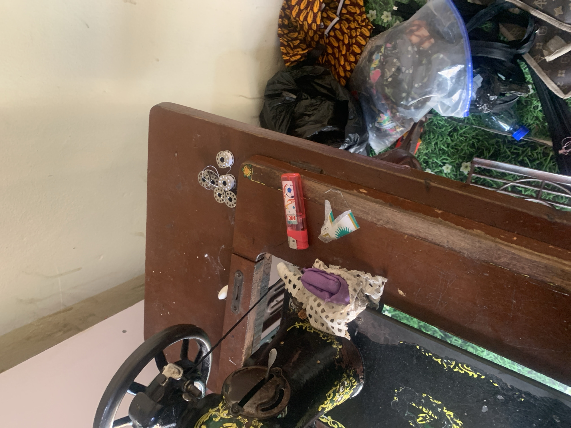 In Jauro Yinu, Nigeria, a close-up shot captures a cluttered sewing area. A vintage sewing machine, predominantly black with gold decorative accents, occupies the left side of the frame. Its handwheel is visible at the top left. The machine sits on a dark brown wooden table, which also displays a collection of sewing spools, some with white thread, and other small metallic items. To the right of the sewing machine, a red lighter with a soccer ball design is present, next to a crumpled white and purple fabric swatch. Further to the right, a black plastic trash bag is prominent, alongside a pile of vibrant, patterned fabric and a clear plastic bag filled with various colorful fabric scraps. A partially visible water bottle with a blue cap rests on the floor, which is covered with green grass and scattered small white flowers. The background consists of a textured, light-colored wall. The overall scene suggests an active, albeit disorganized, crafting space.